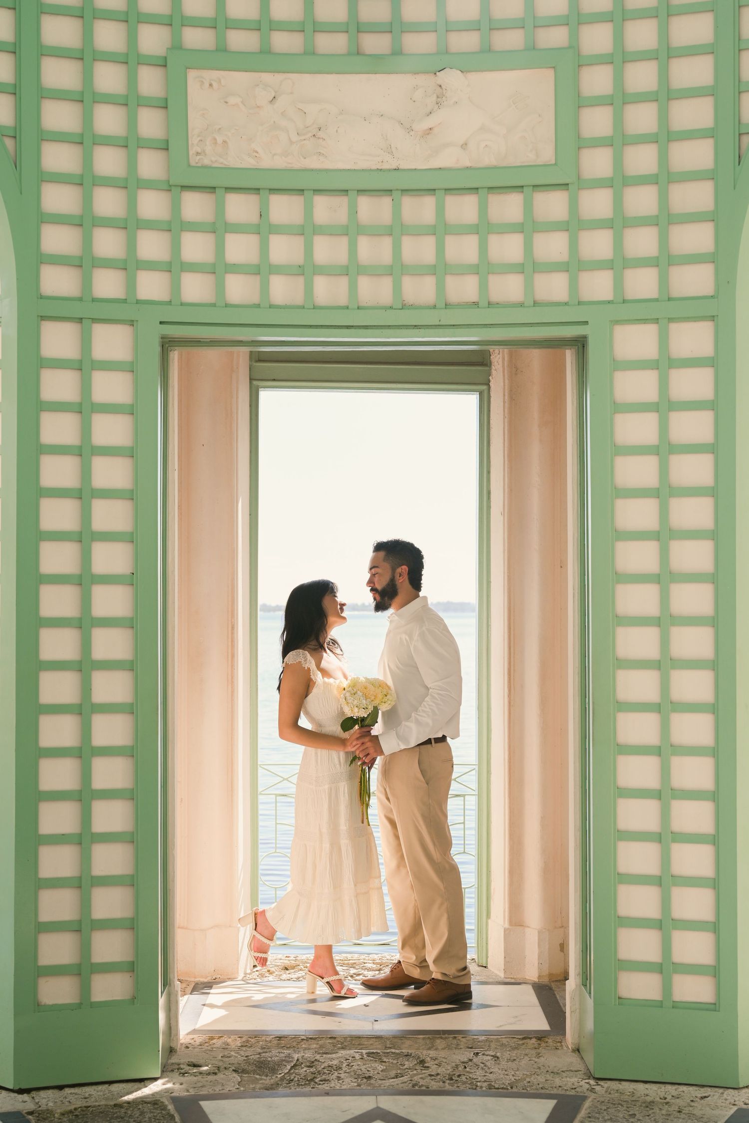 A couple in white outfits stand by a window in a mint green room with lattice wall details.