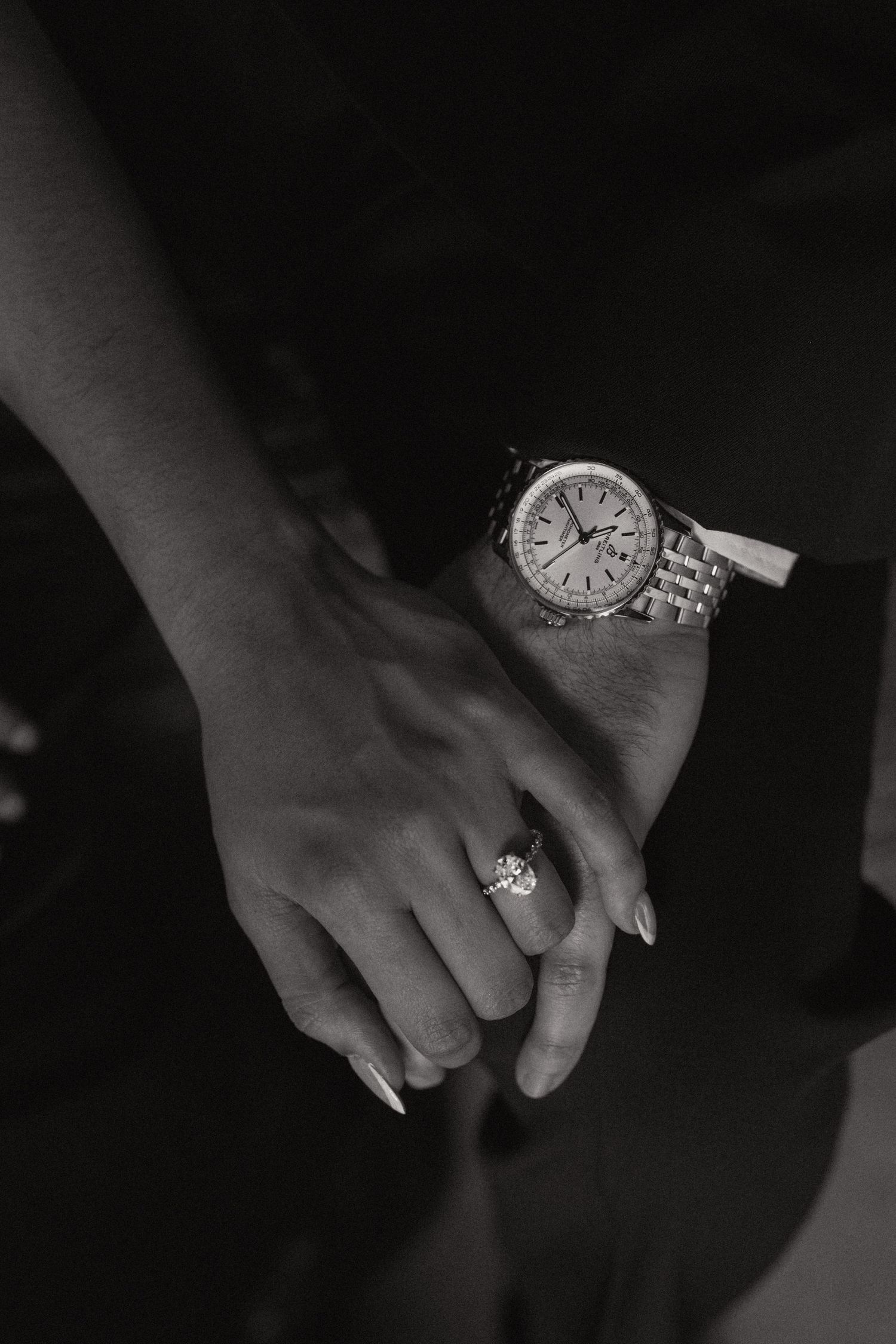 Black and white close-up of a couple holding hands, showing a diamond engagement ring and a classic wristwatch.
