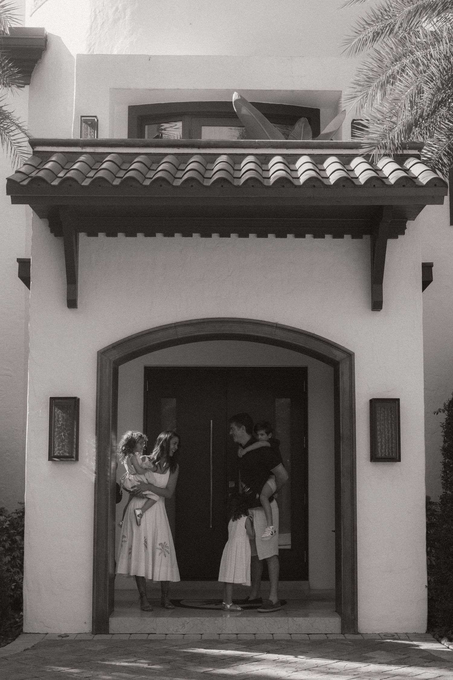 Black and white photo of a family of four standing in the arched doorway of a Mediterranean-style home with a tile roof and palm fronds.