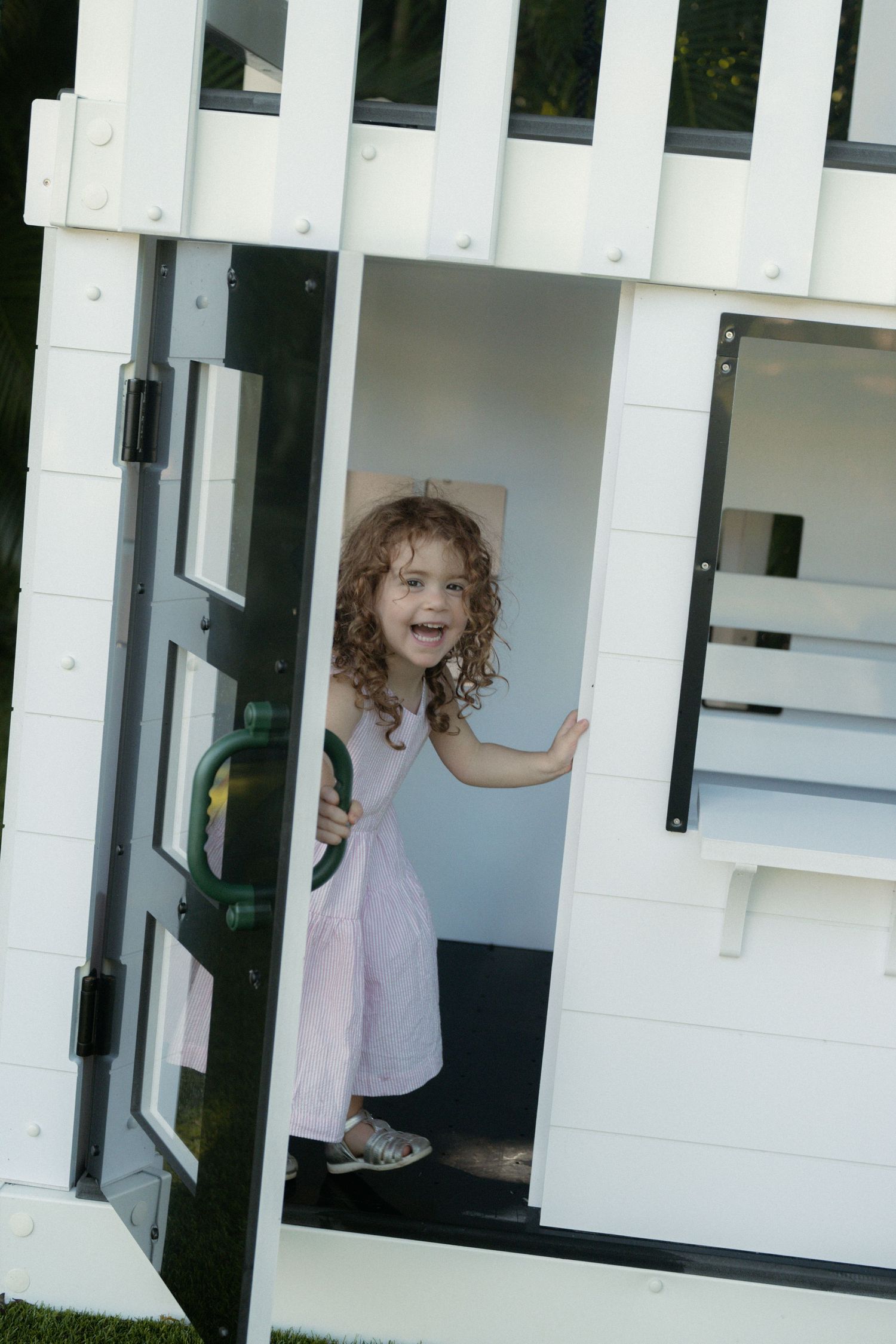 Smiling little girl with curly hair in a pink dress peeks out the open door of a white wooden outdoor playhouse.