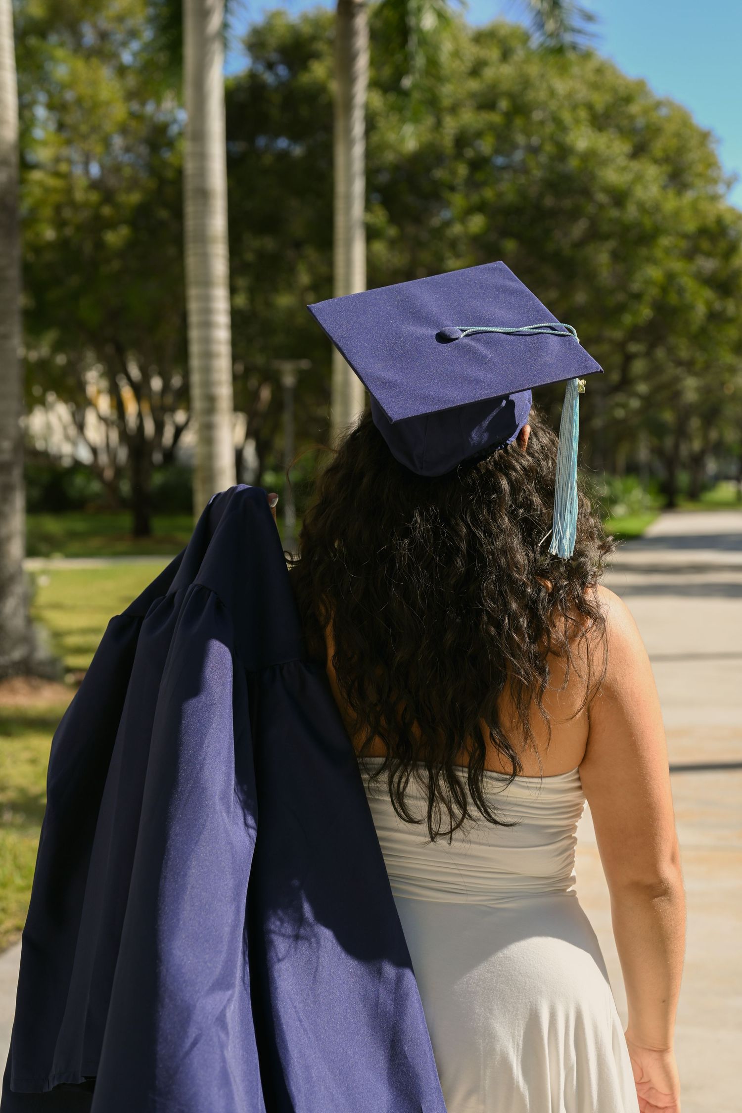 Graduate wearing navy cap and gown stands on outdoor campus pathway.