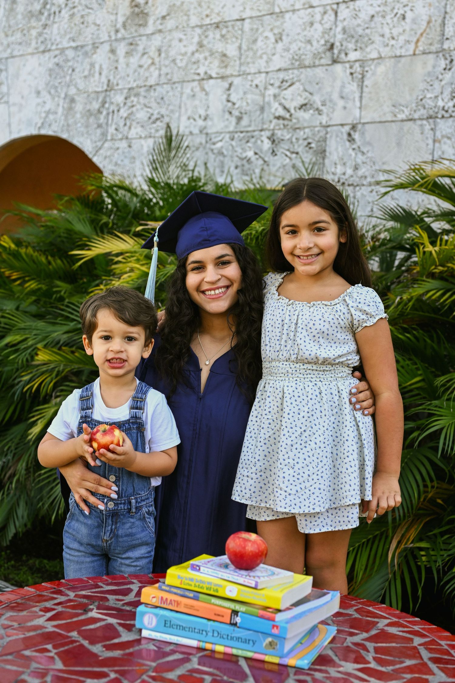 Graduate in navy regalia celebrates with family on red tile patio with educational books.