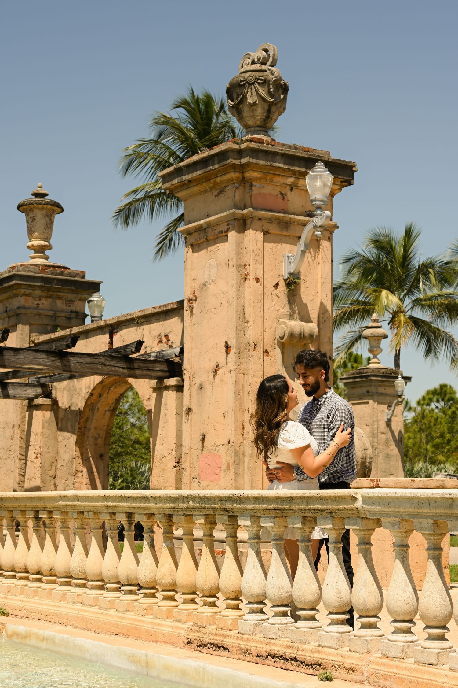 A couple embraces by ornate stone pillars with palm trees swaying in the background.