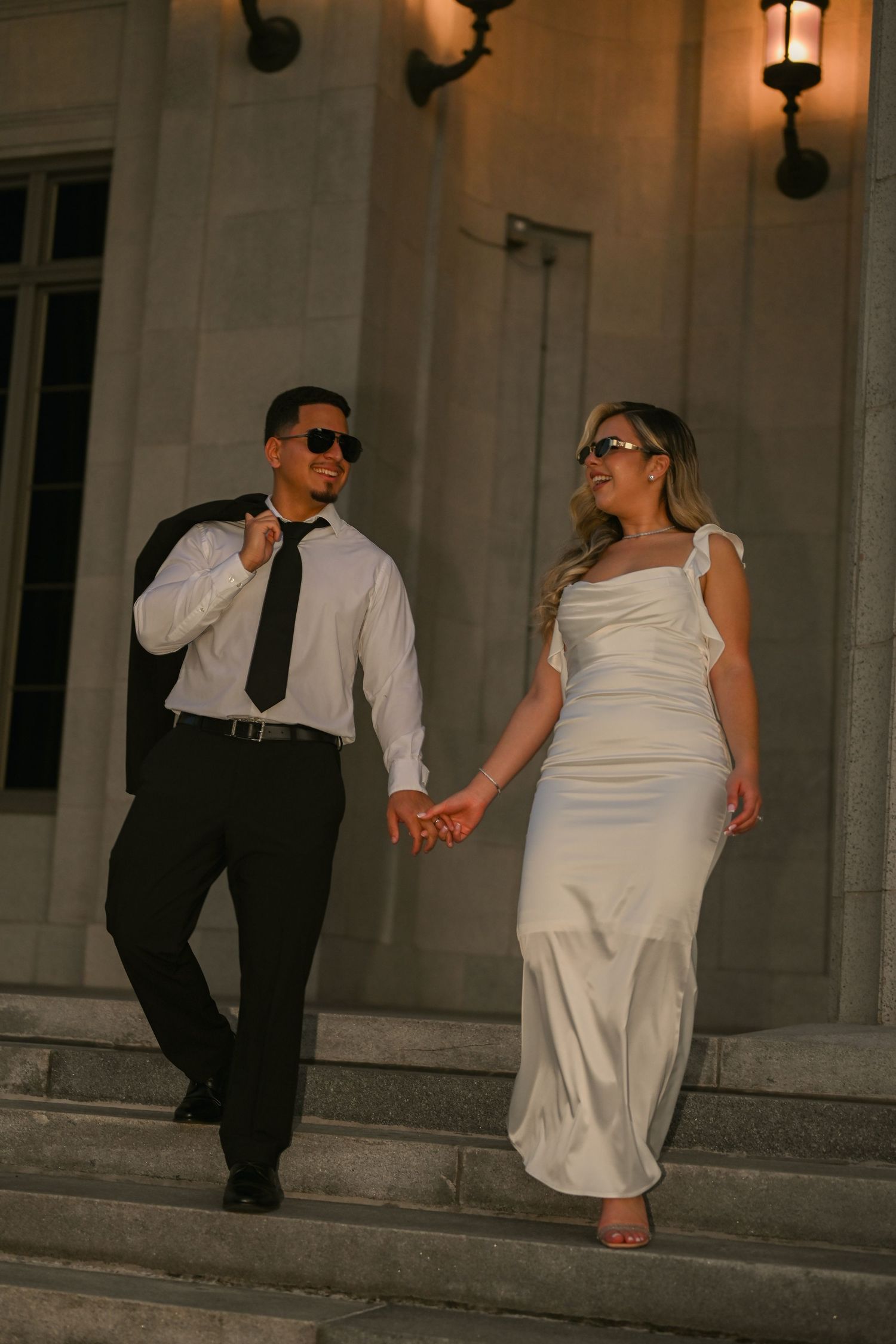 Wedding couple in sunglasses dancing playfully on stone steps at dusk.