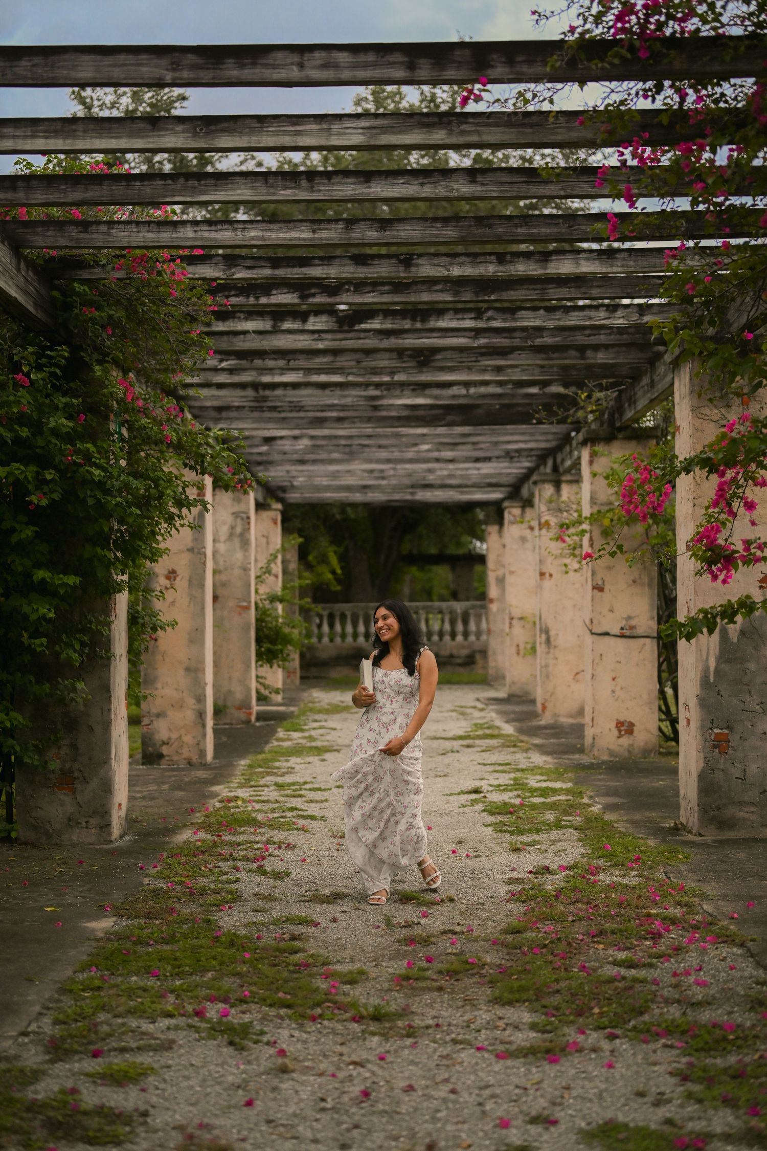 A floral-dressed figure moves through a stone colonnade with scattered pink flowers.