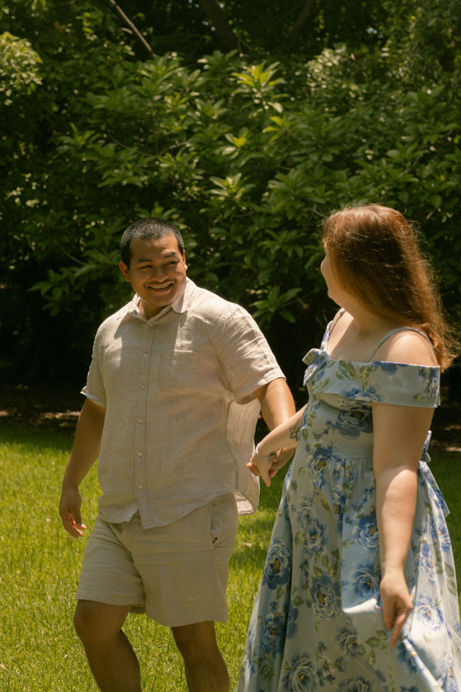A smiling couple holds hands while walking together on a sunny green lawn surrounded by lush trees.