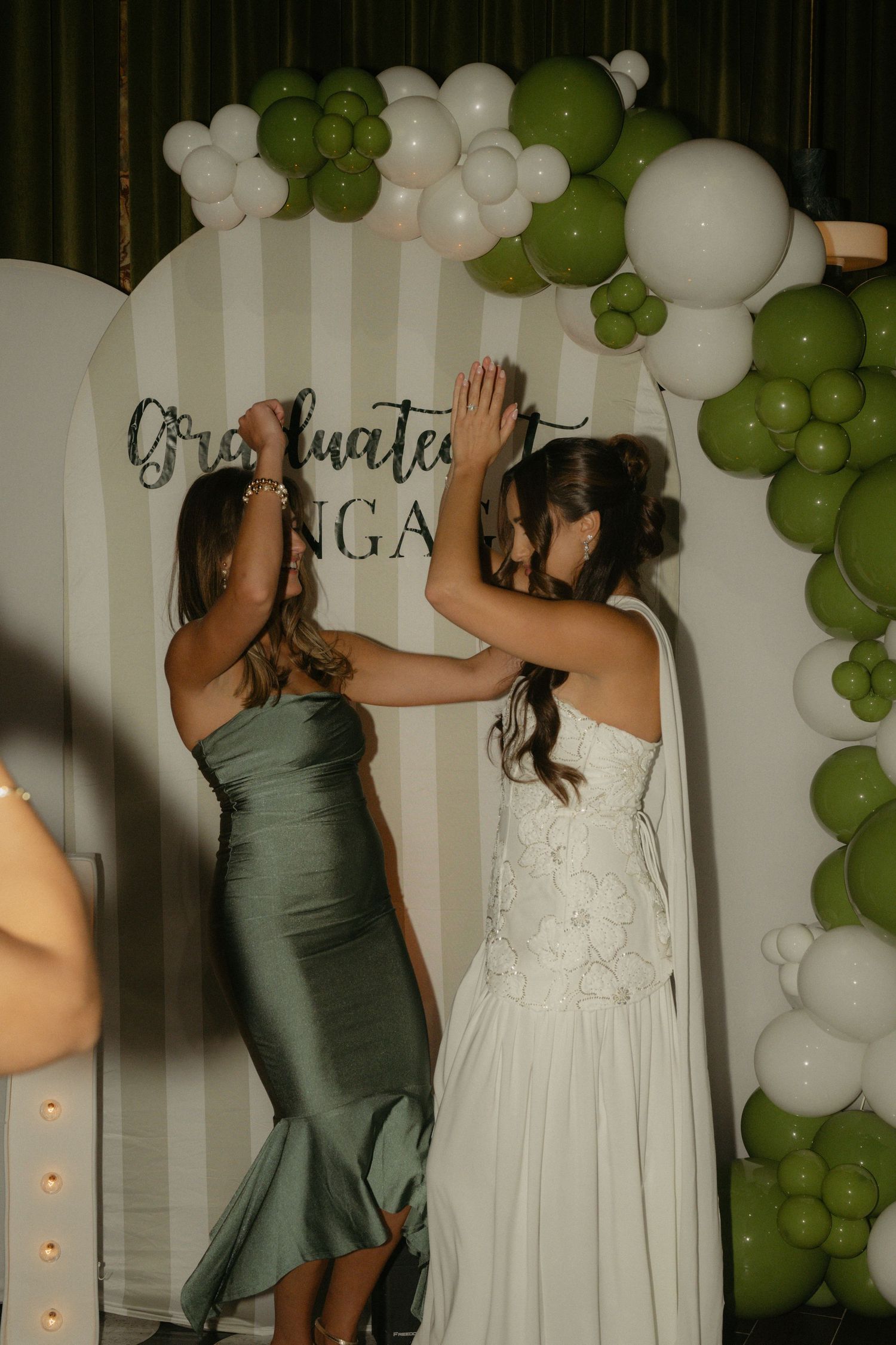 Two women posing at a graduation party backdrop with green and white balloon arch decor.