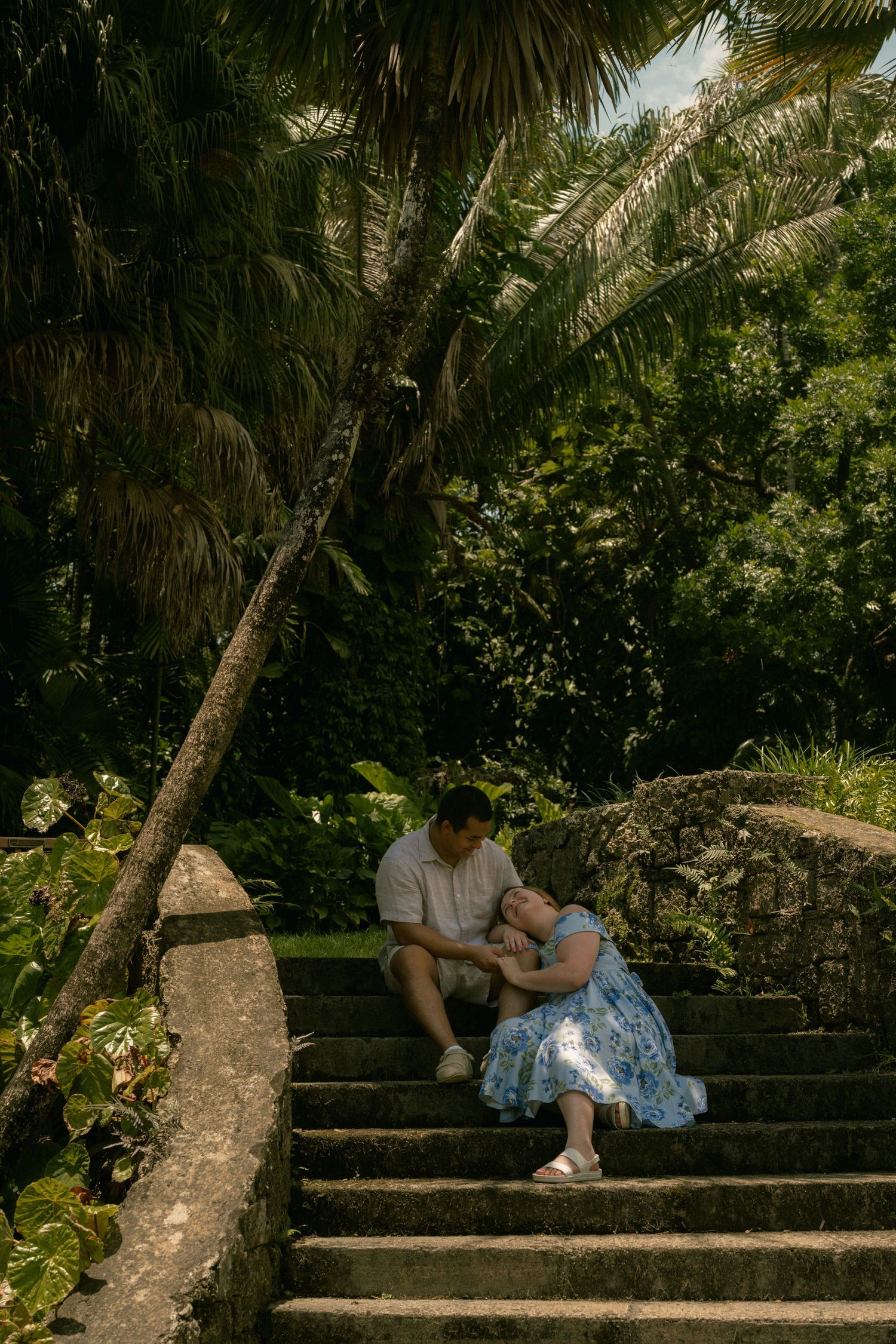 Couple sitting on stone steps surrounded by lush tropical palm trees and greenery in an outdoor garden setting.