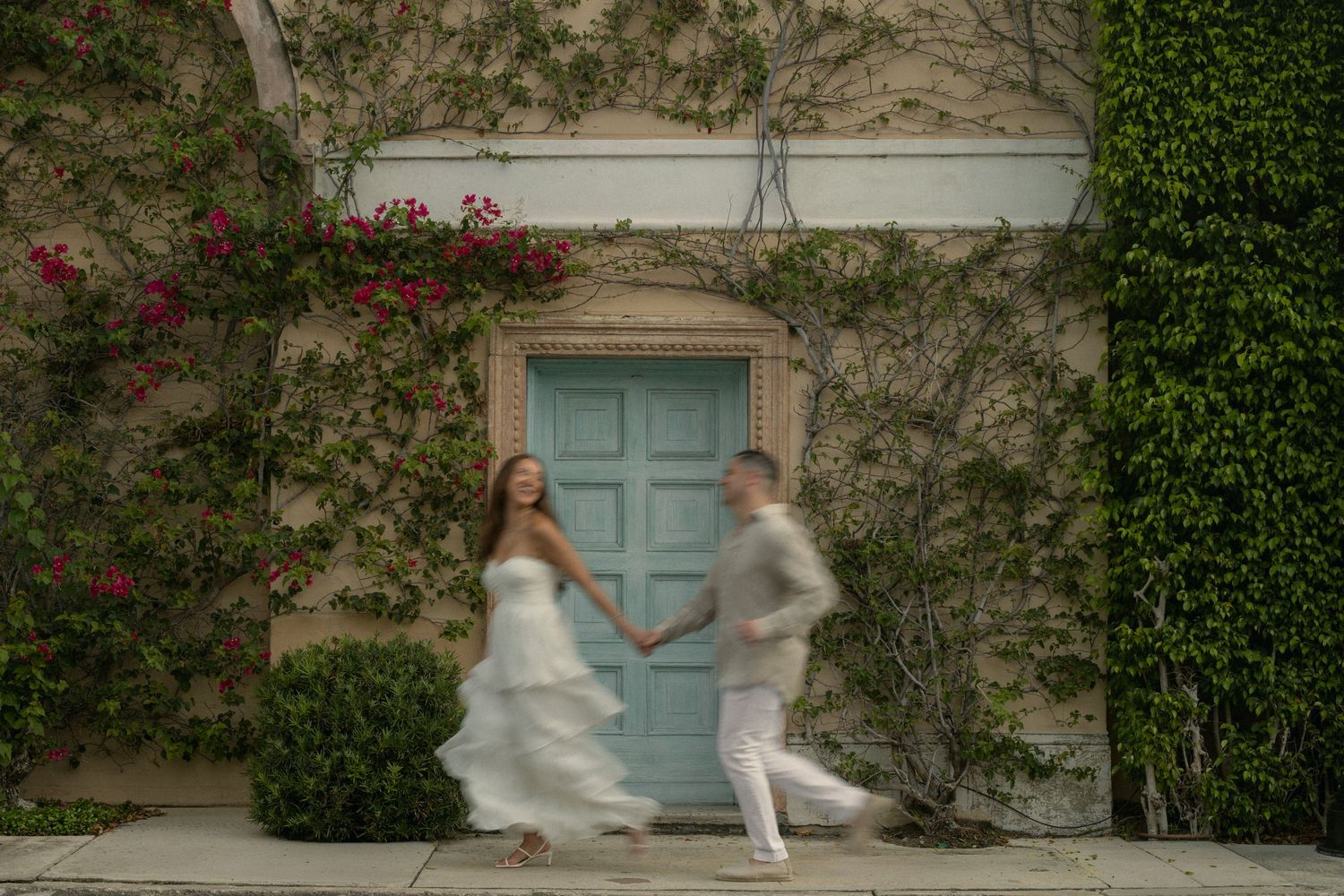Blurred motion shot of bride and groom holding hands walking past ivy-covered wall with blue door.