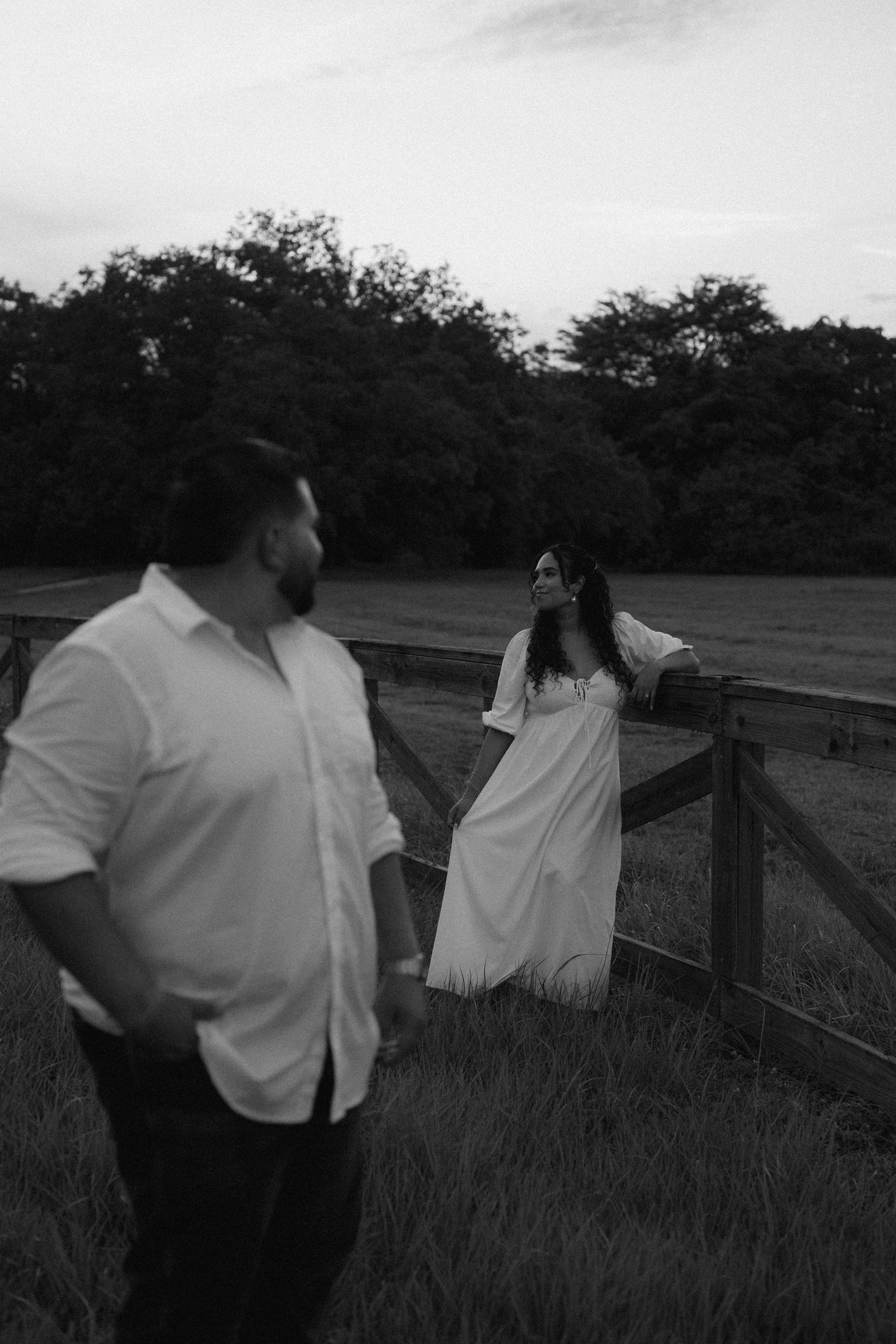 Black and white photo of a couple near a wooden fence in a field, woman in white dress gazes up while man stands nearby.