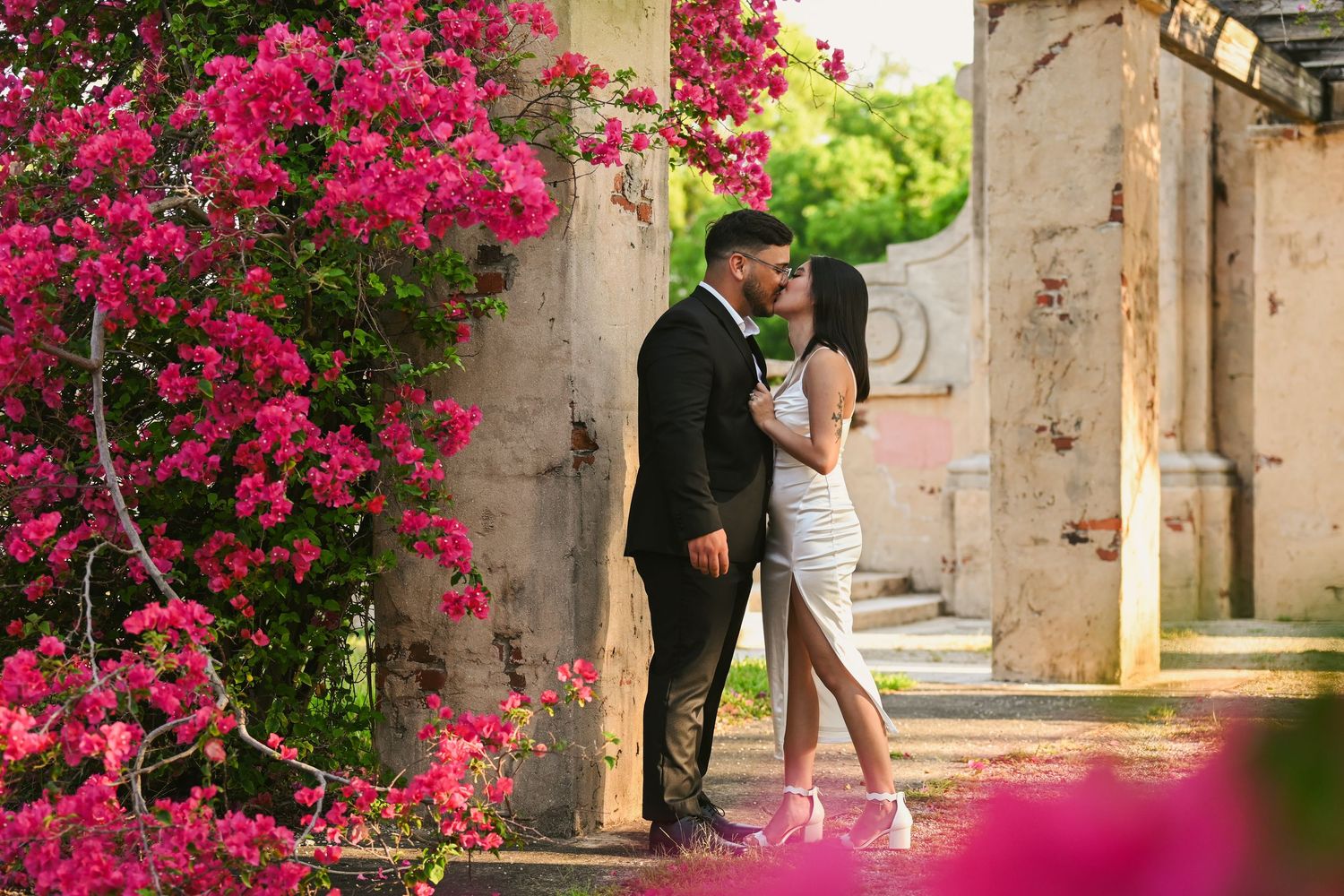Couple sharing an intimate moment beneath pink flowering vines in an outdoor garden setting.