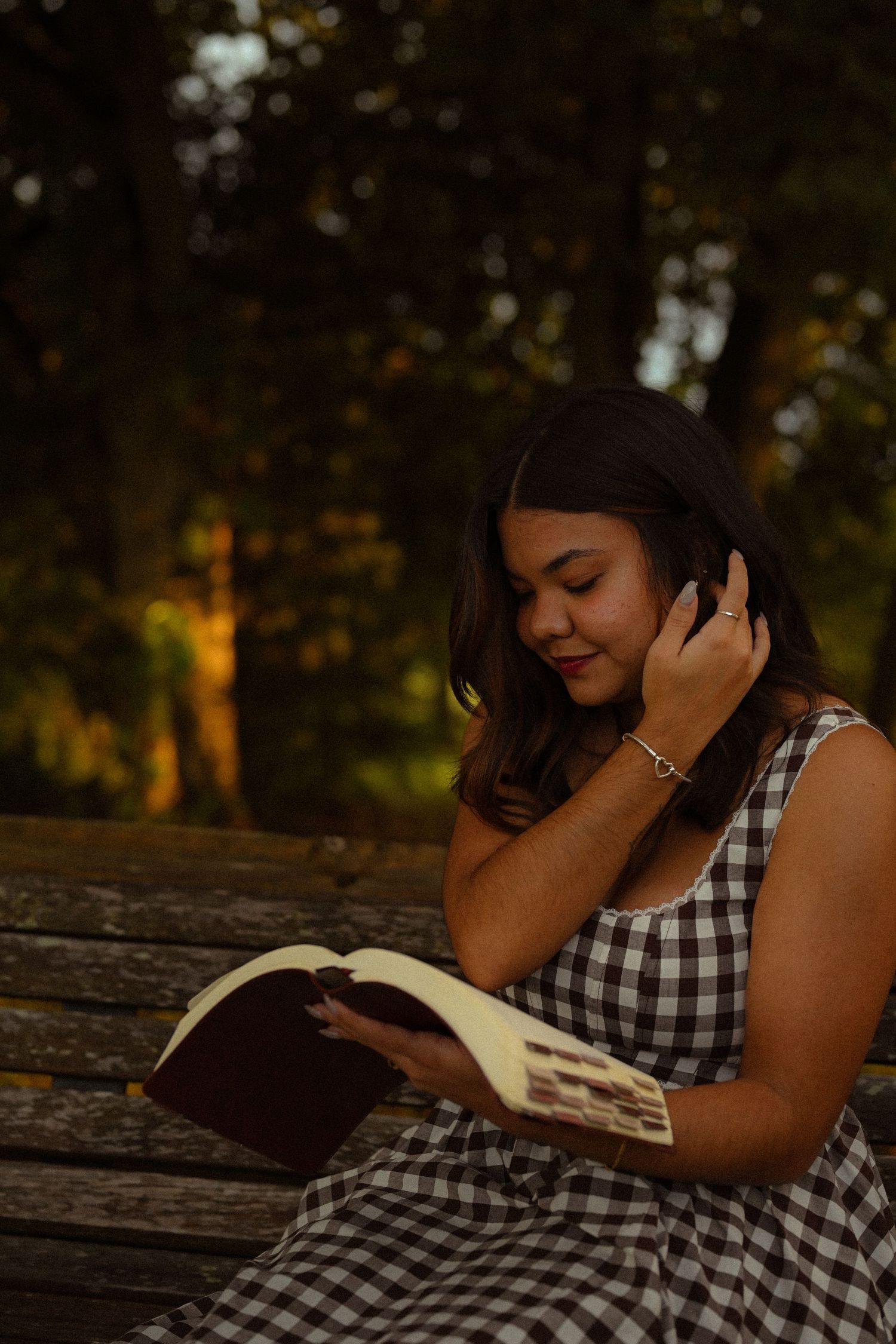 A person in a gingham dress reads quietly on a weathered wooden bench in a park setting.