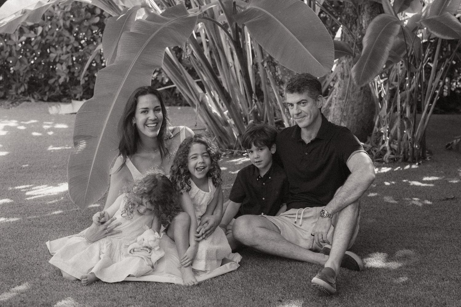 Black and white family portrait of a smiling woman, man, two boys and a young girl sitting together on the ground among tropical plants.