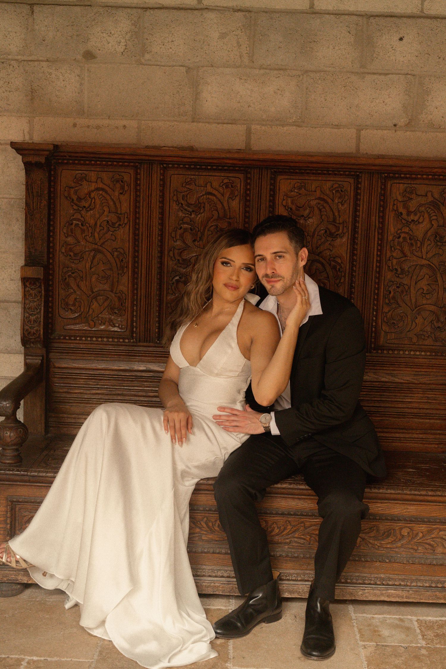Bride in a white satin halter gown and groom in a dark suit sit close on an ornately carved wooden bench against a stone wall.