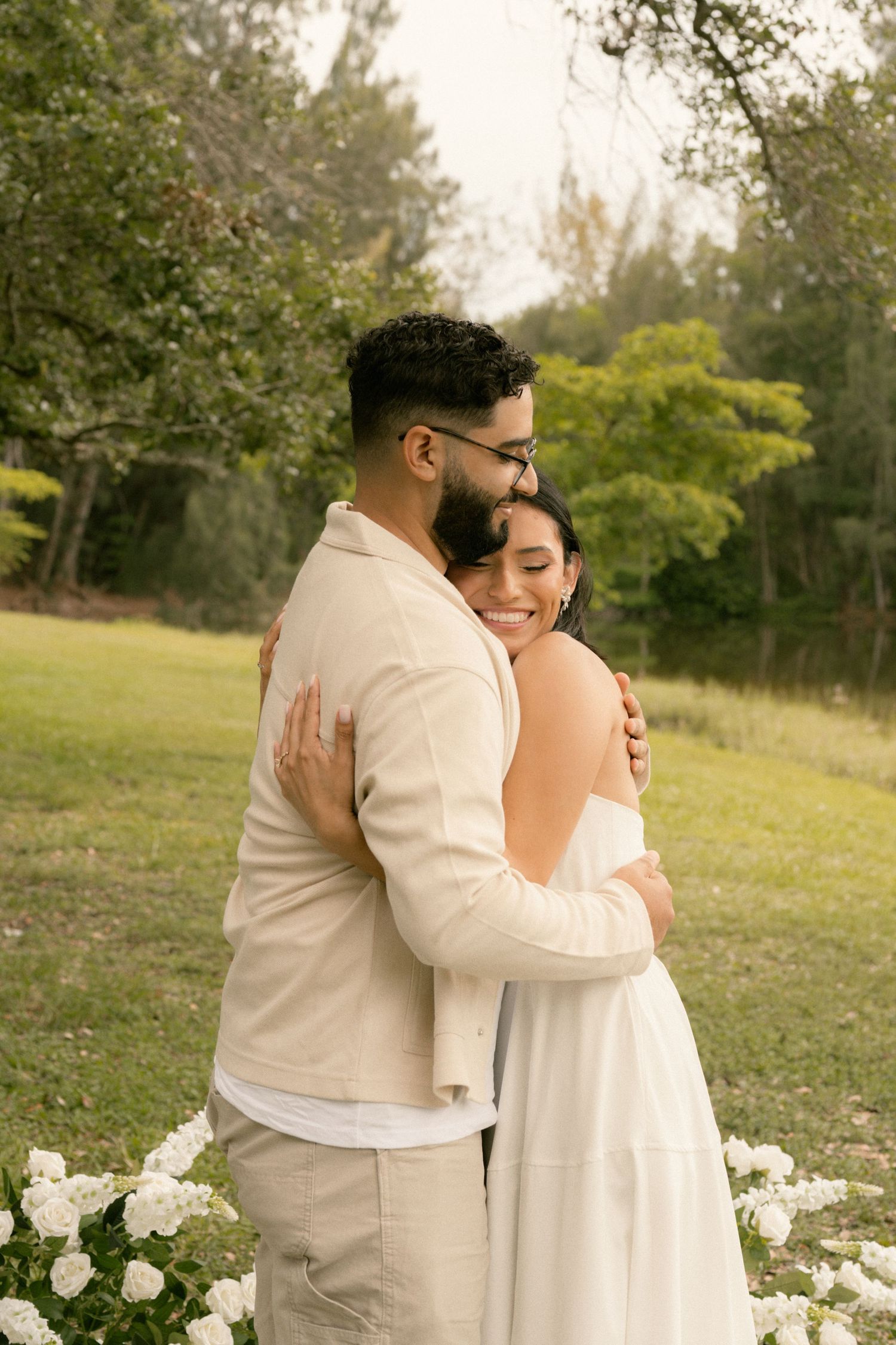 Couple in white and beige outfits embracing outdoors near white flowers and lush green trees.