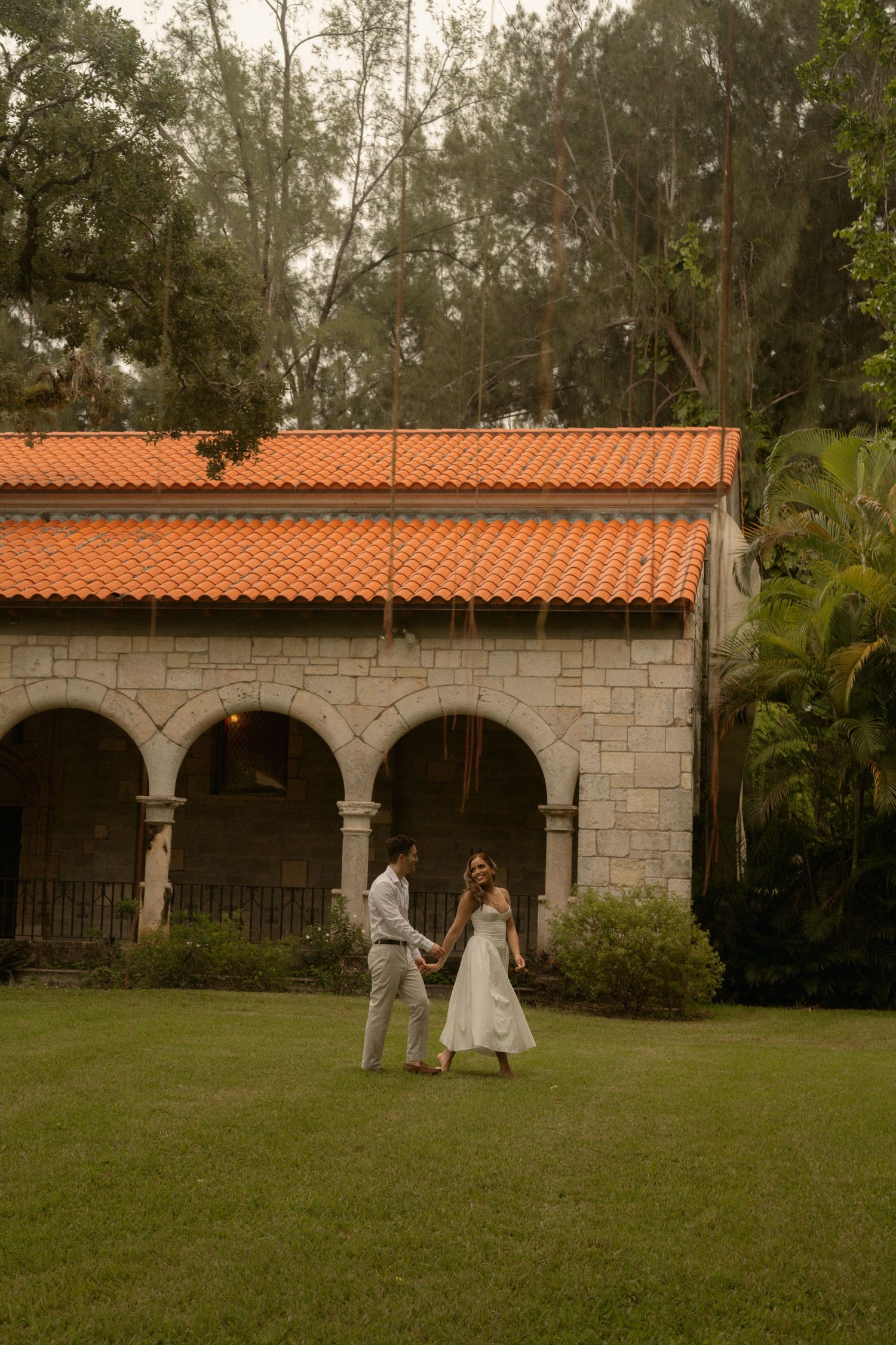 Couple in white outfits holding hands and walking across a green lawn toward a historic stone building with arches and a terracotta roof.
