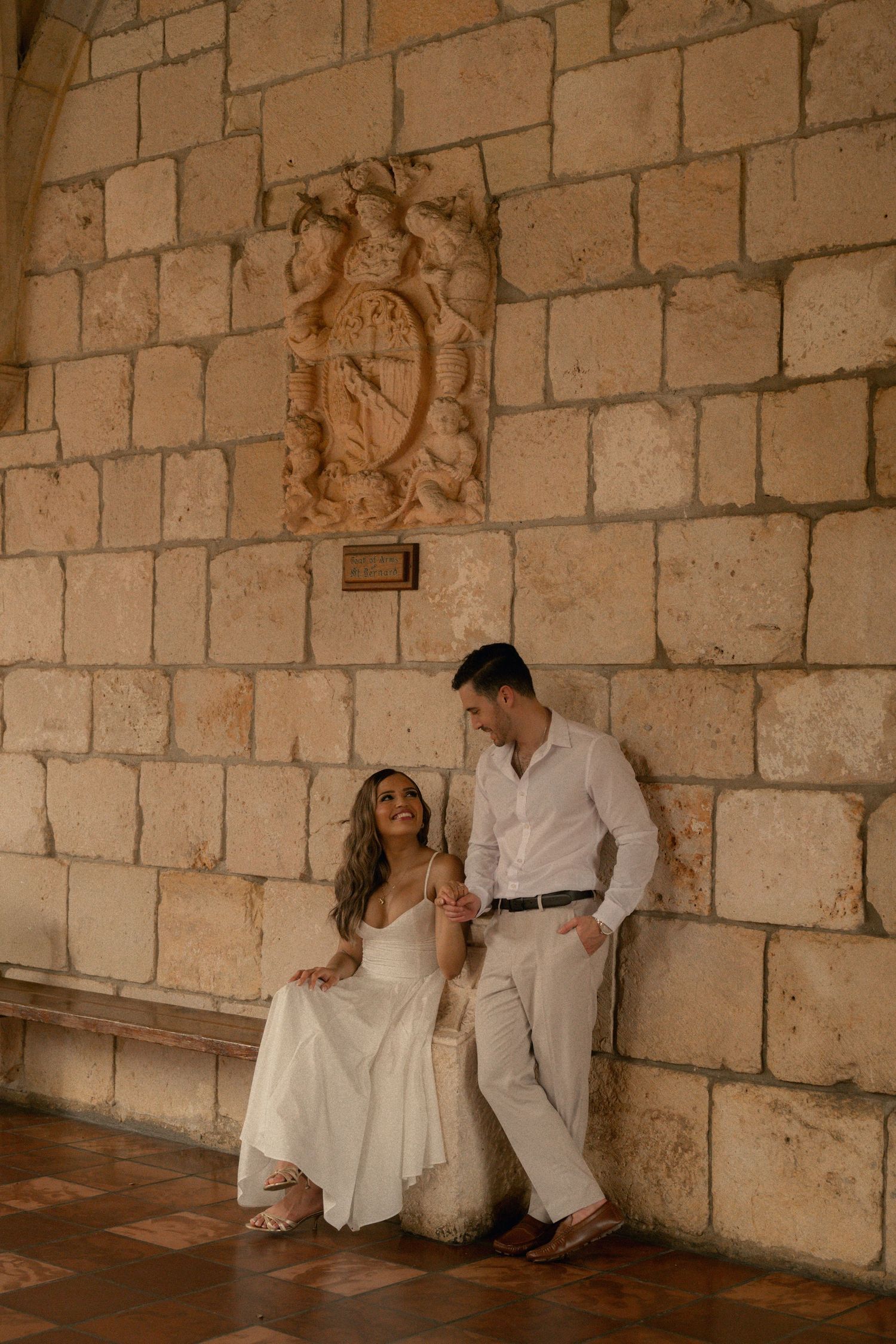 Bride sits on a stone bench as the groom leans beside her against a historic stone wall with a carved coat of arms.
