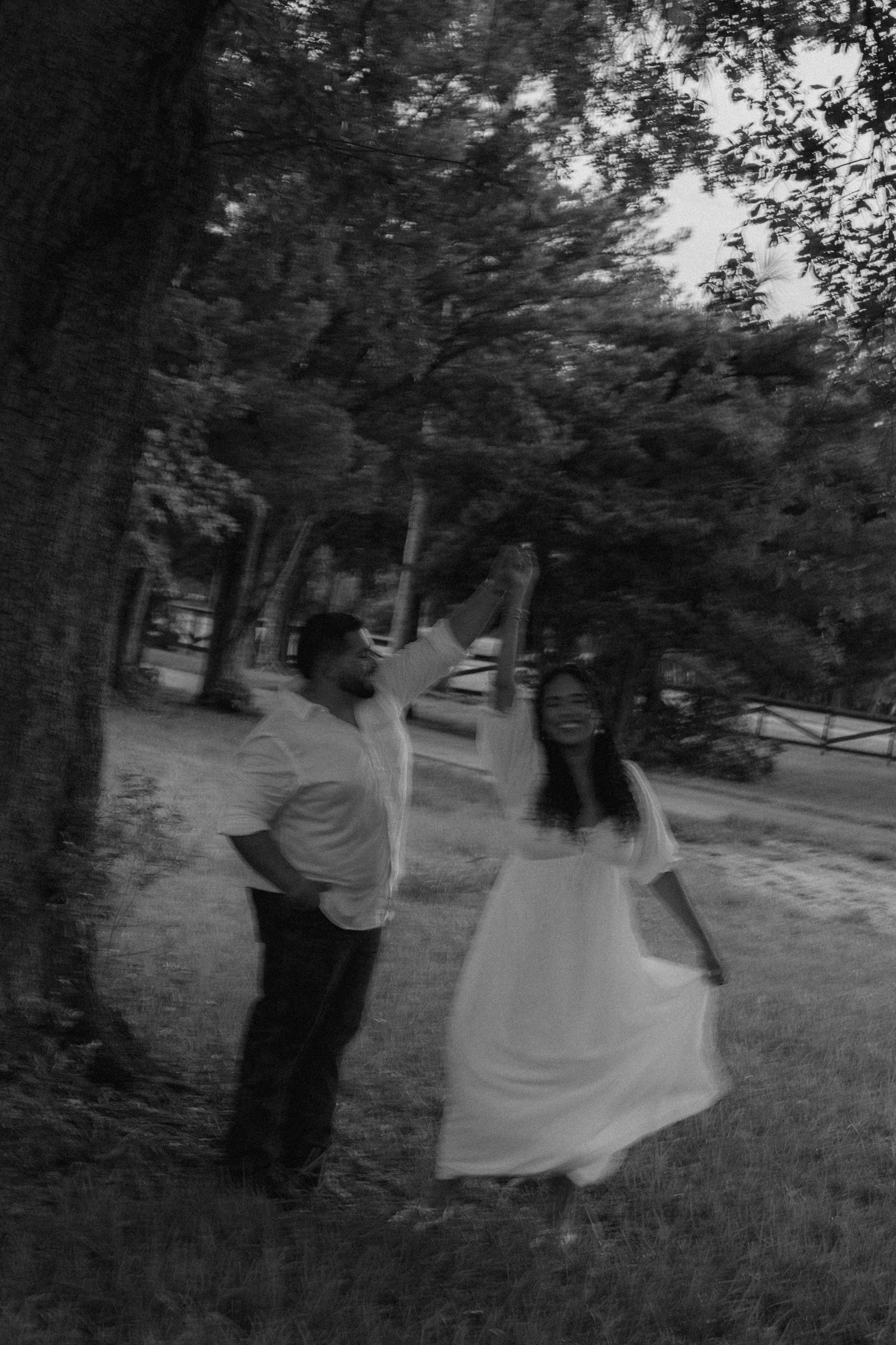 Black and white photo of a couple dancing outdoors near trees, woman in flowing white dress, man in casual attire.
