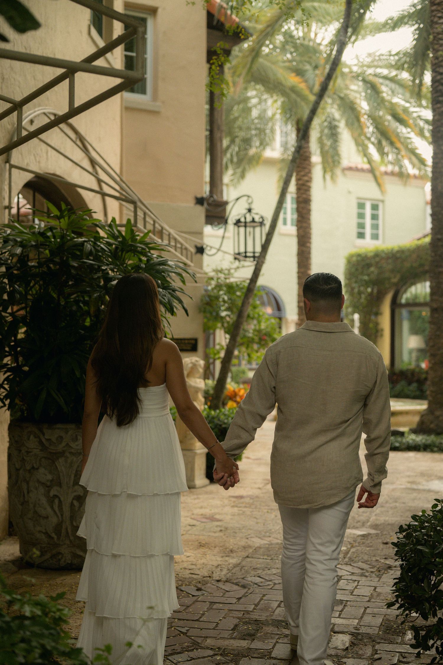 A couple holds hands walking through a lush tropical courtyard with palm trees and Mediterranean architecture.