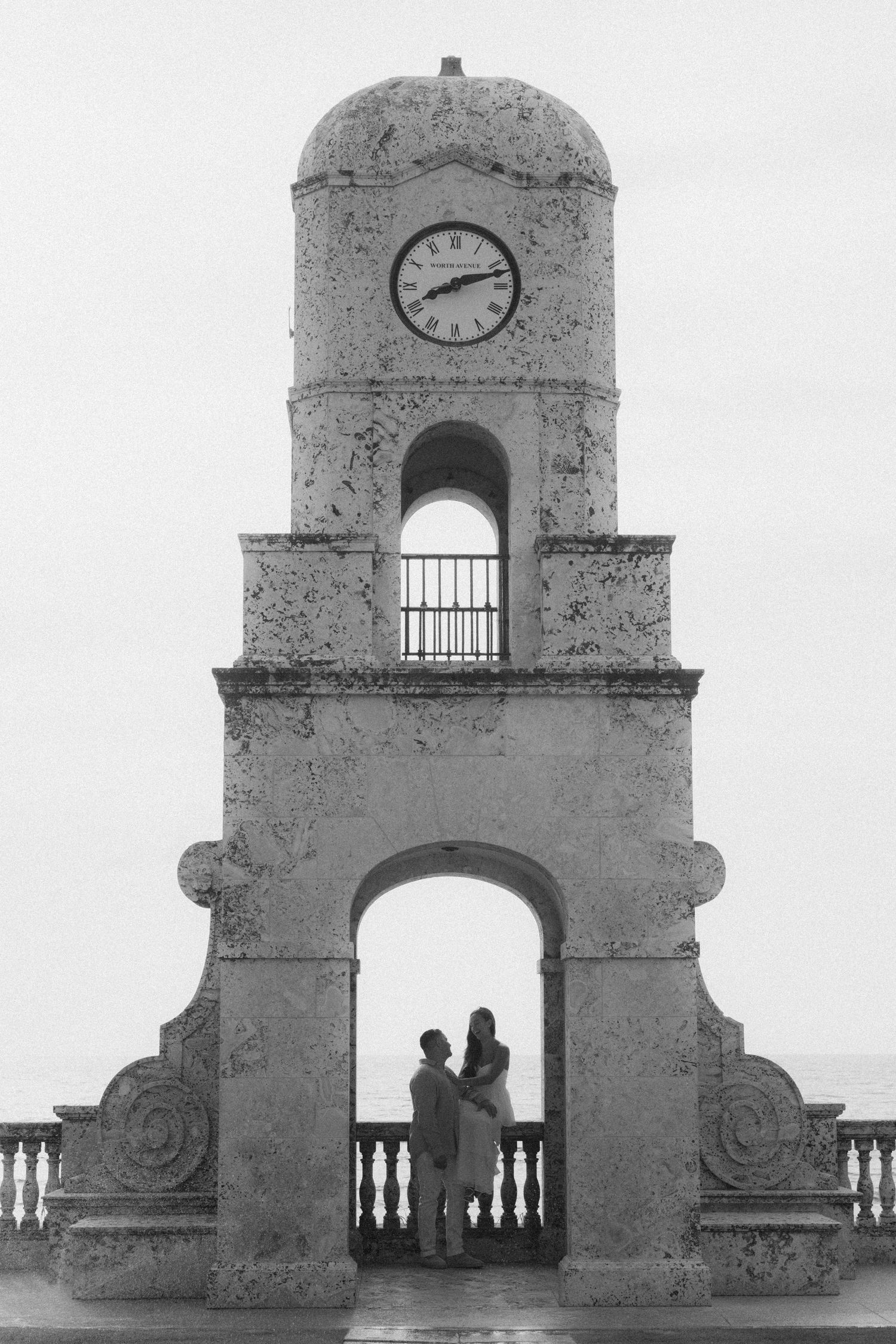 Couple silhouetted under historic clock tower arch in black and white romantic photo.