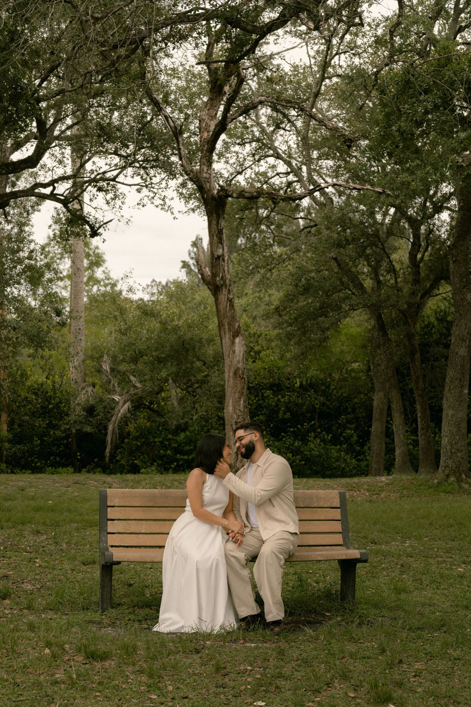 Bride and groom sitting on a wooden park bench surrounded by lush green trees in an outdoor wedding photo.