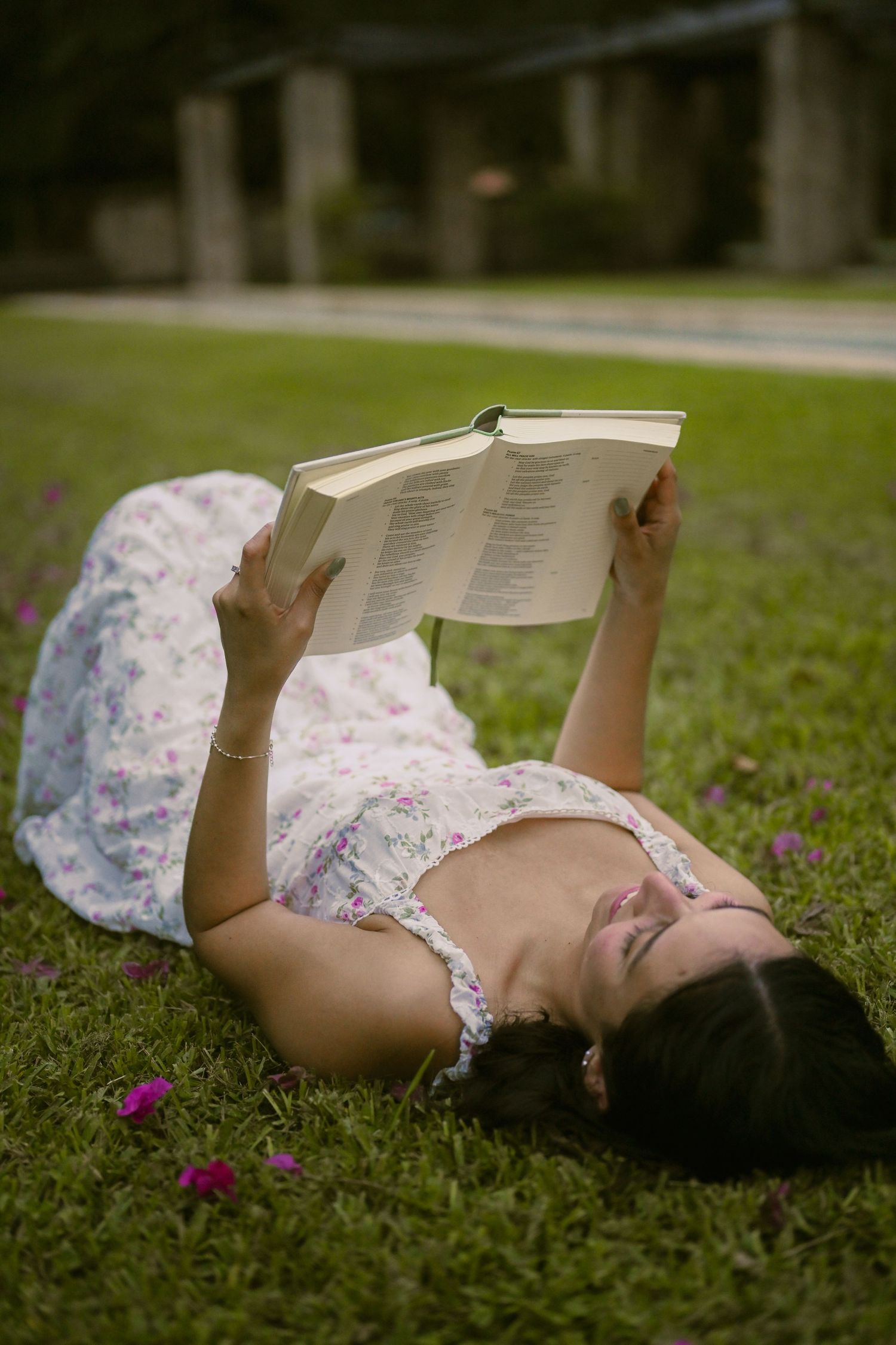 A reader relaxes on grass holding a book overhead while wearing a floral dress.