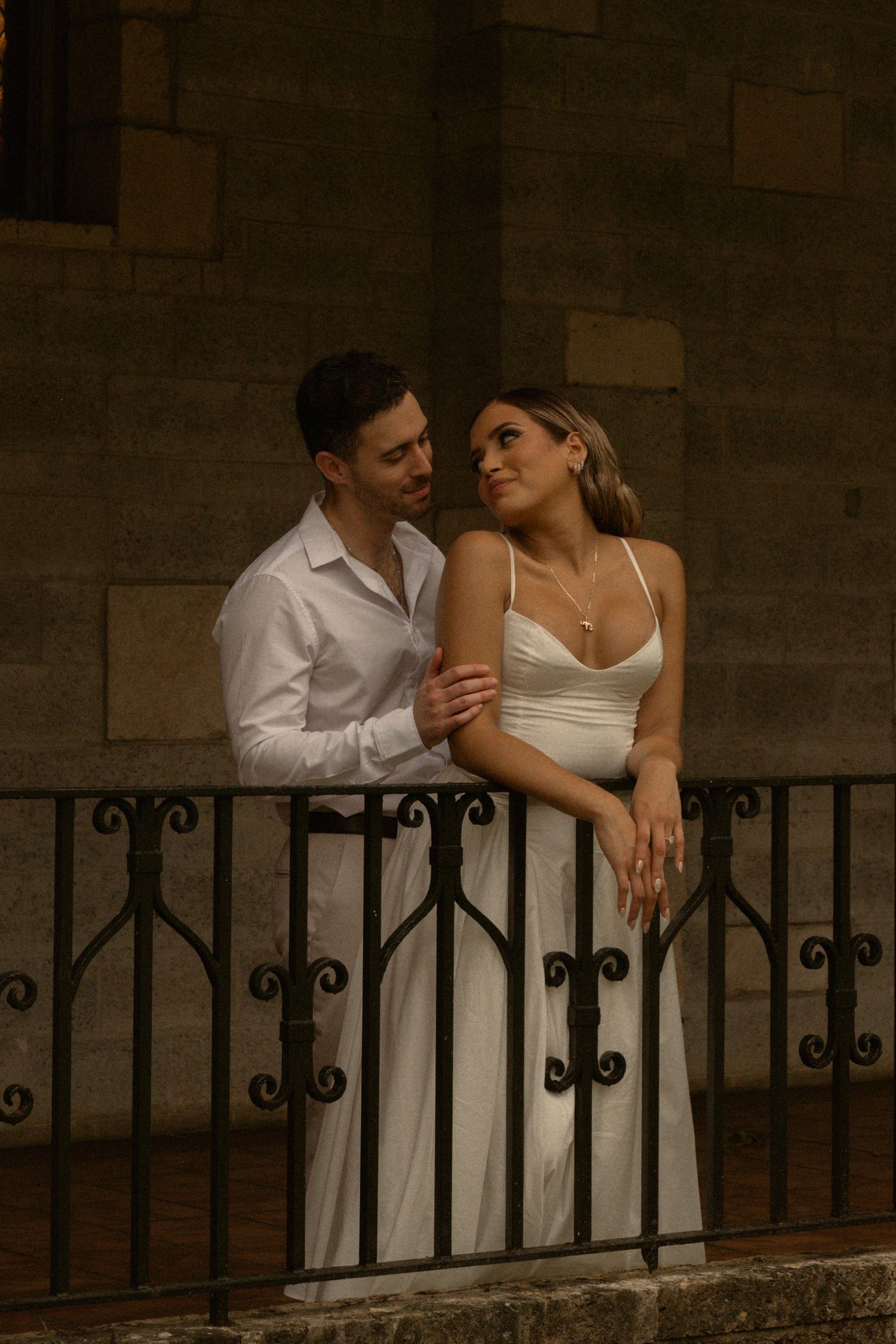 Couple in white outfits embrace on a stone balcony with a wrought iron railing during an elegant engagement photo session.