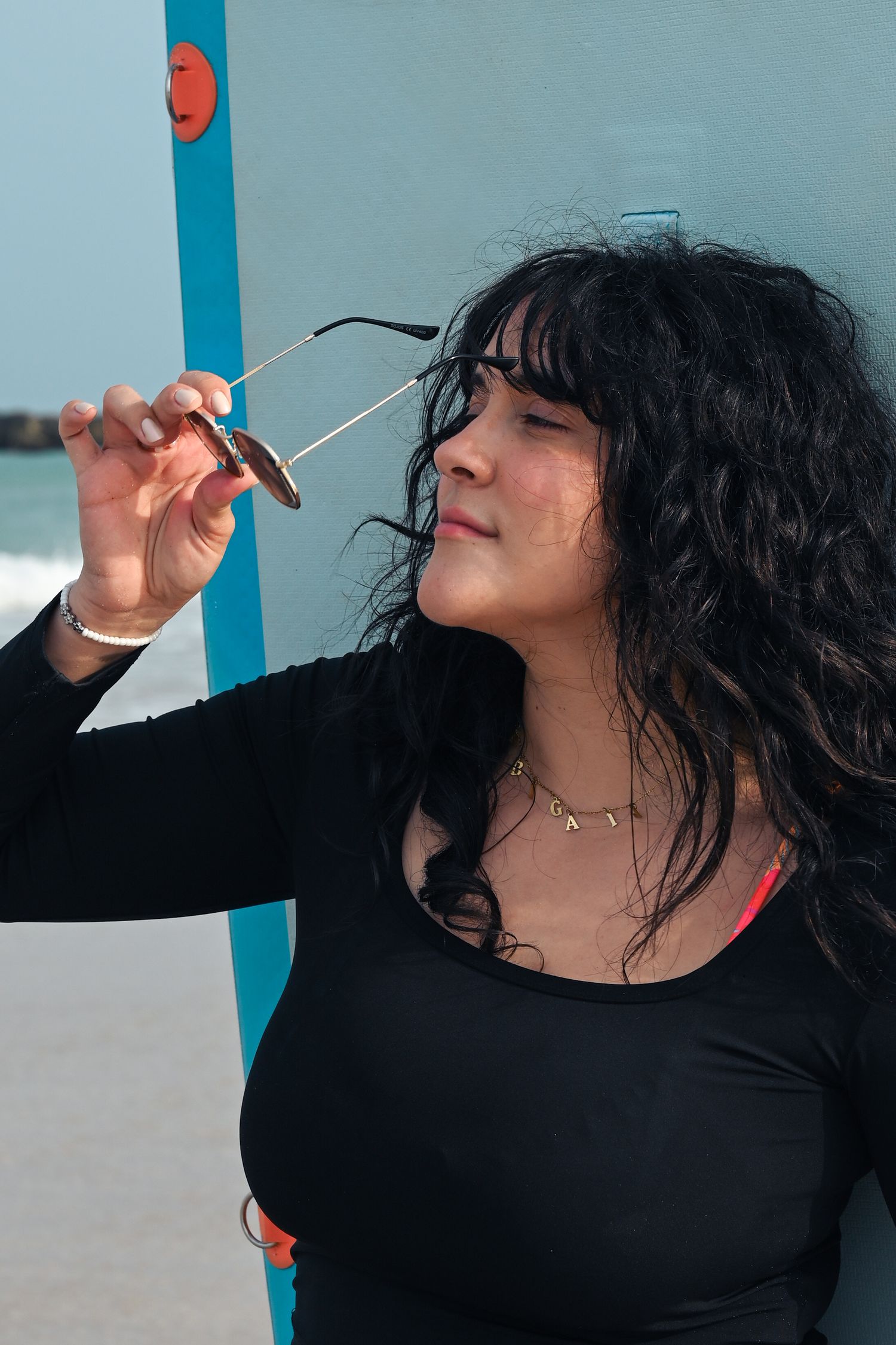 Person in black long sleeve top poses expressively against blue wall at the beach.