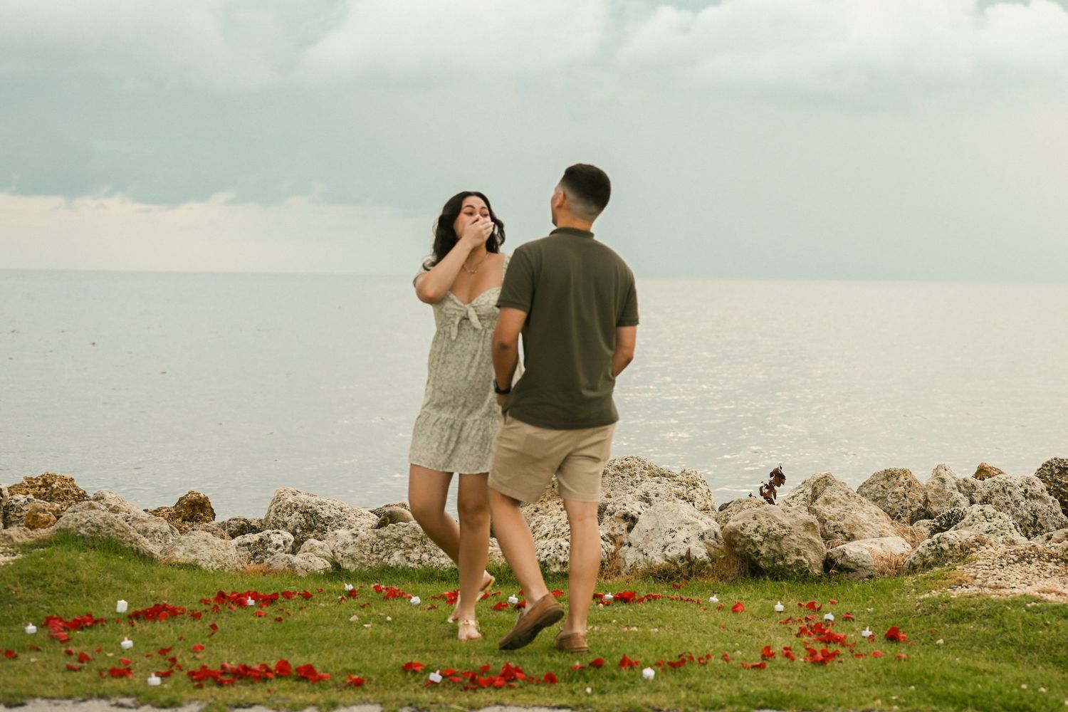A couple stands on a rocky oceanside cliff with scattered red flowers at sunset.
