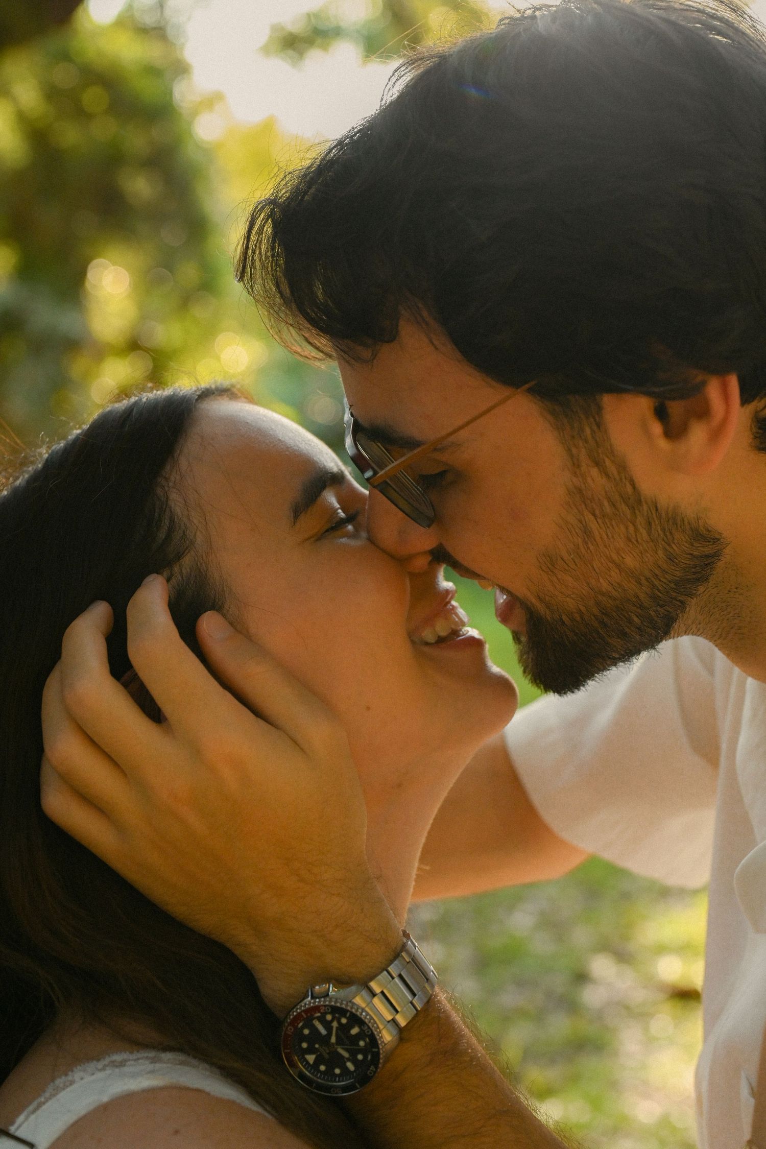 A romantic couple sharing tender kisses in a sunlit outdoor setting with natural bokeh effect in the background.