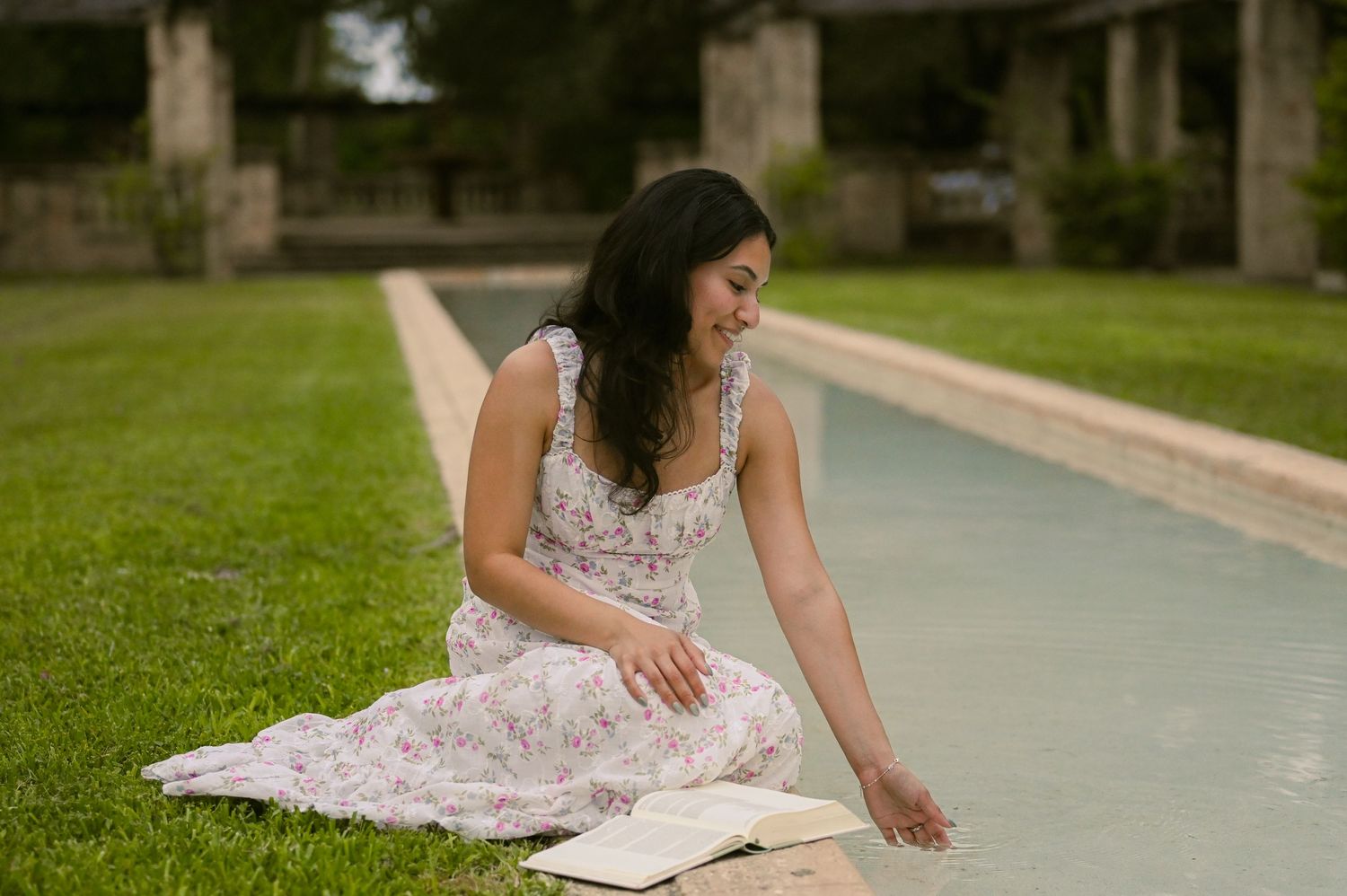 A person in a floral sundress sits reading on a path surrounded by grass.