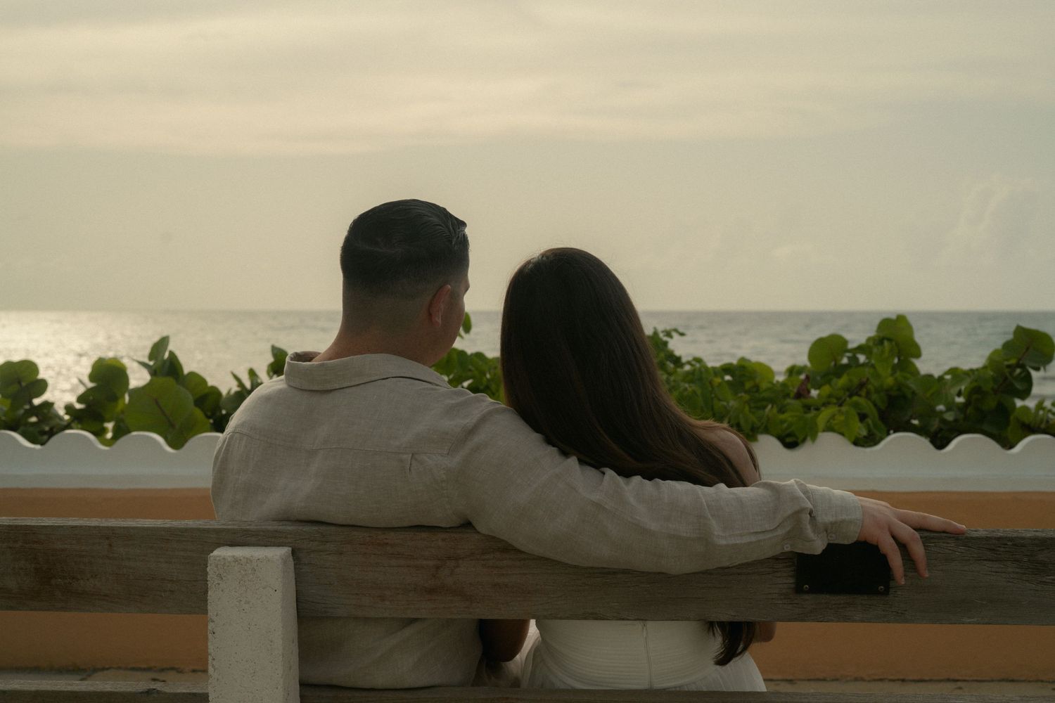 Couple sitting on a bench overlooking the ocean at sunset, sharing a romantic moment by the sea.