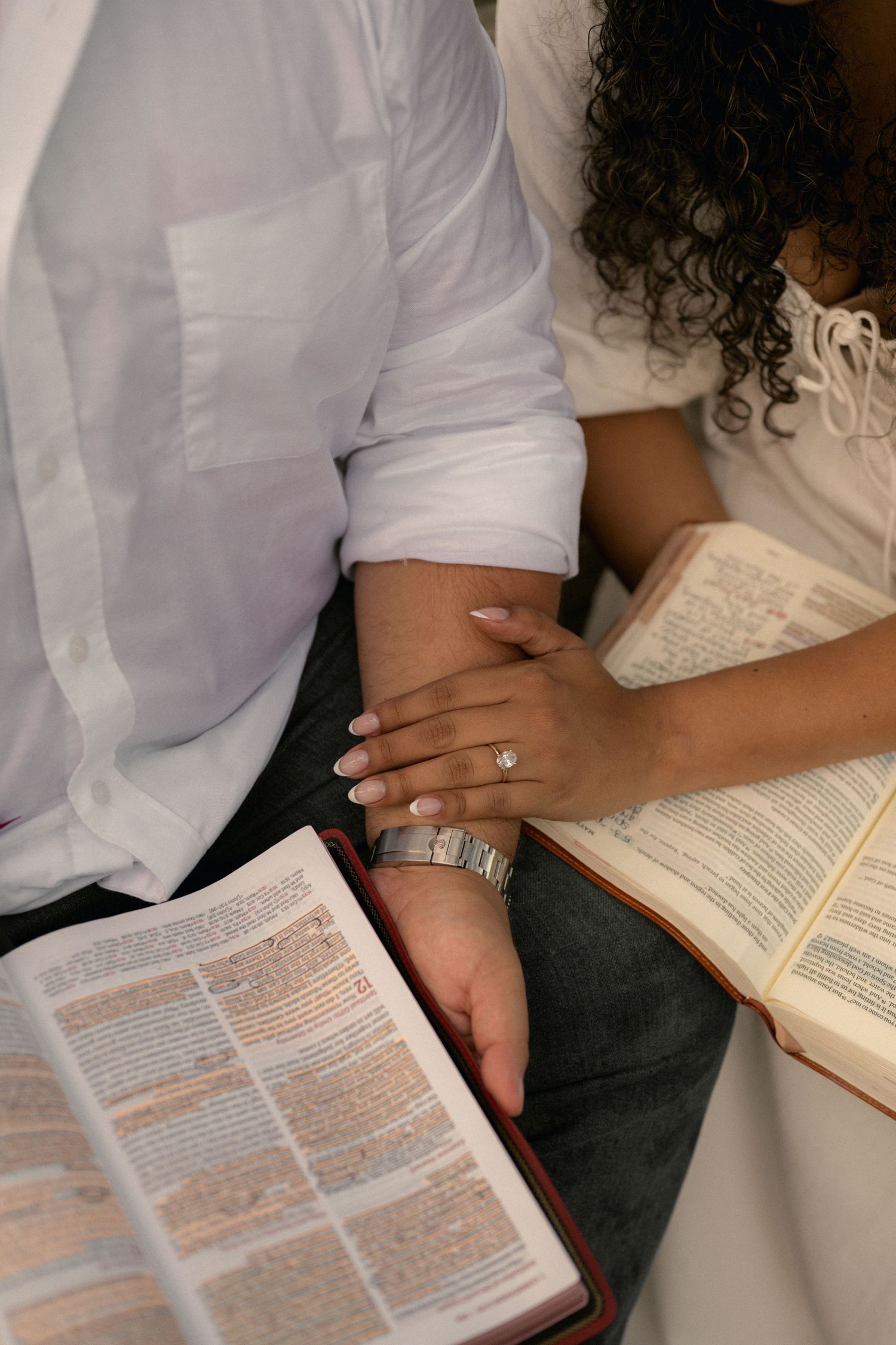 Couple sitting together holding hands with open Bibles, woman wearing engagement ring, faith and love concept.