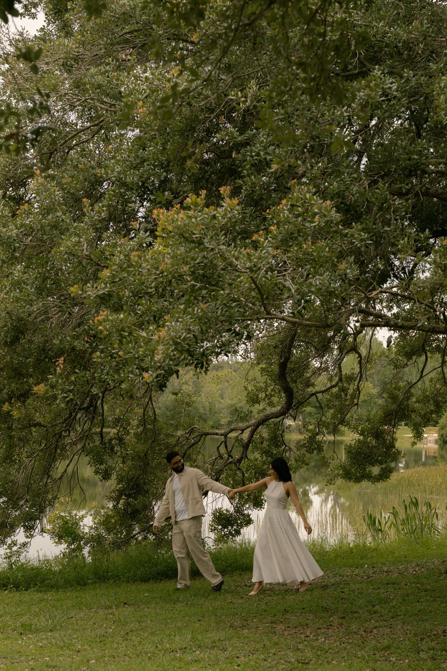 Couple in white outfits holding hands walking near a serene lake under large green trees in a lush park.