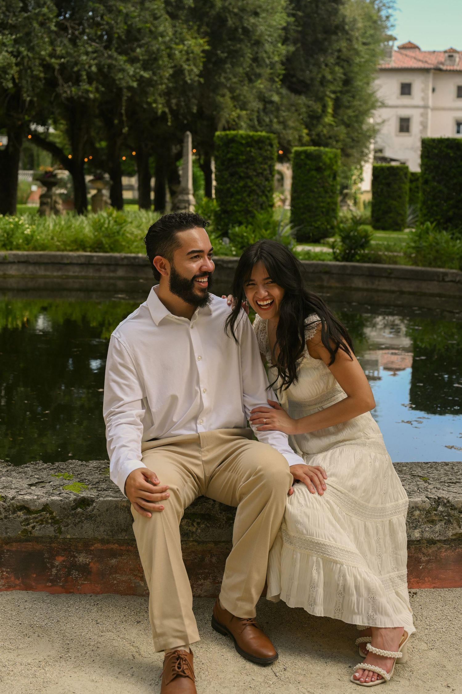 A couple shares a romantic moment sitting together by a reflective pond in a garden setting.