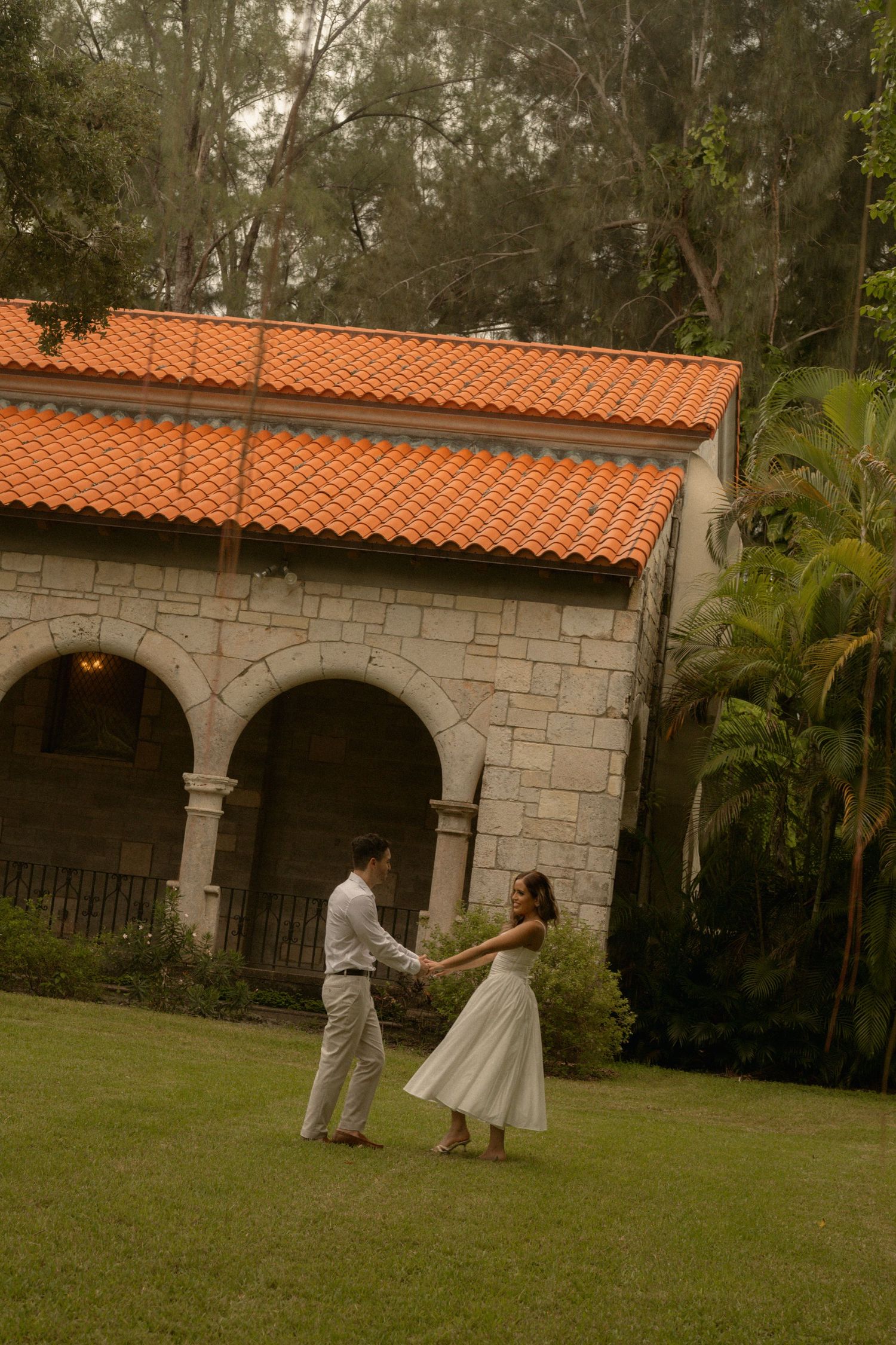 Couple in wedding attire holds hands and dances on a green lawn in front of a stone building with an orange terracotta tile roof.