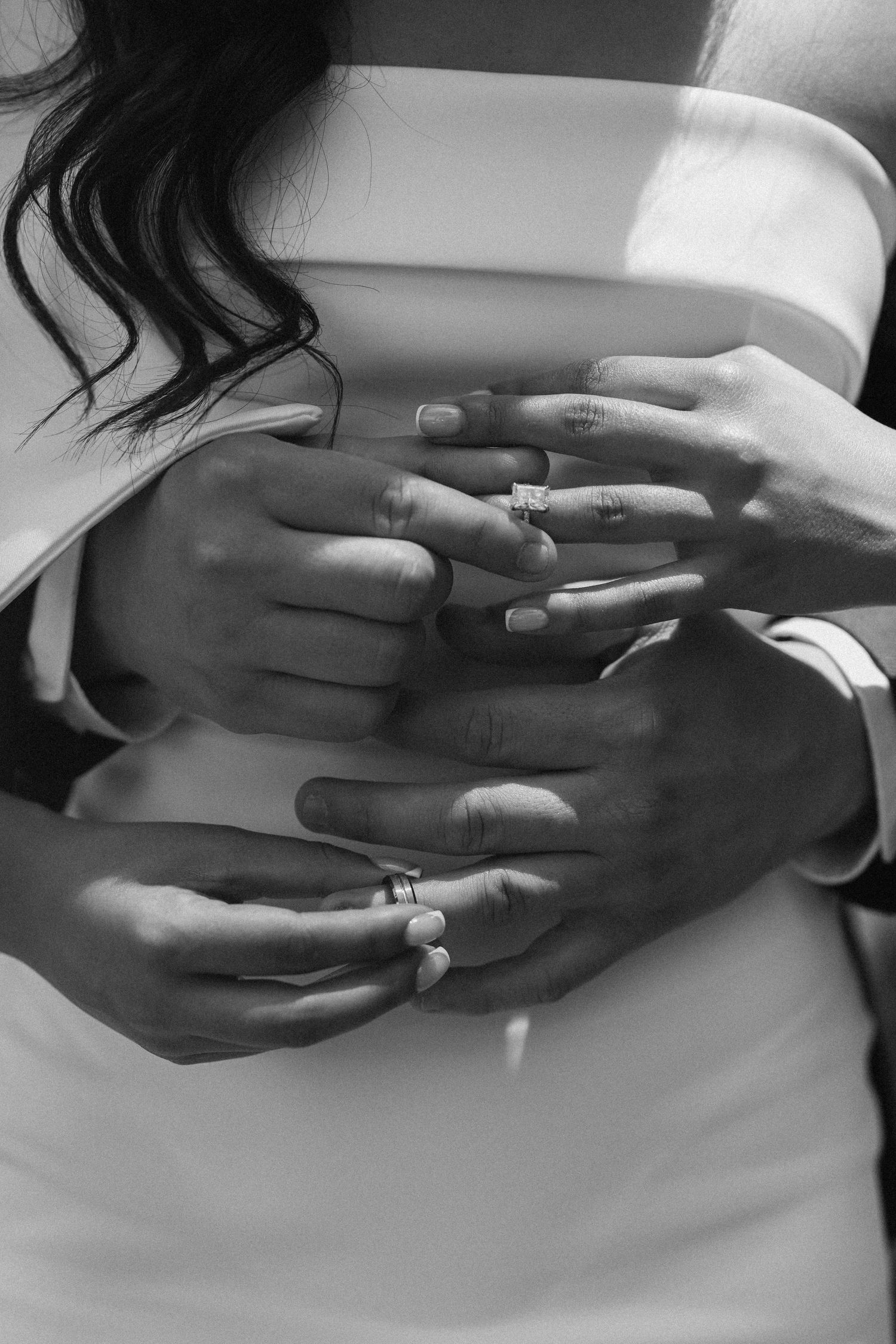 Black and white photo of newlyweds embracing, showcasing wedding and engagement rings on their hands.