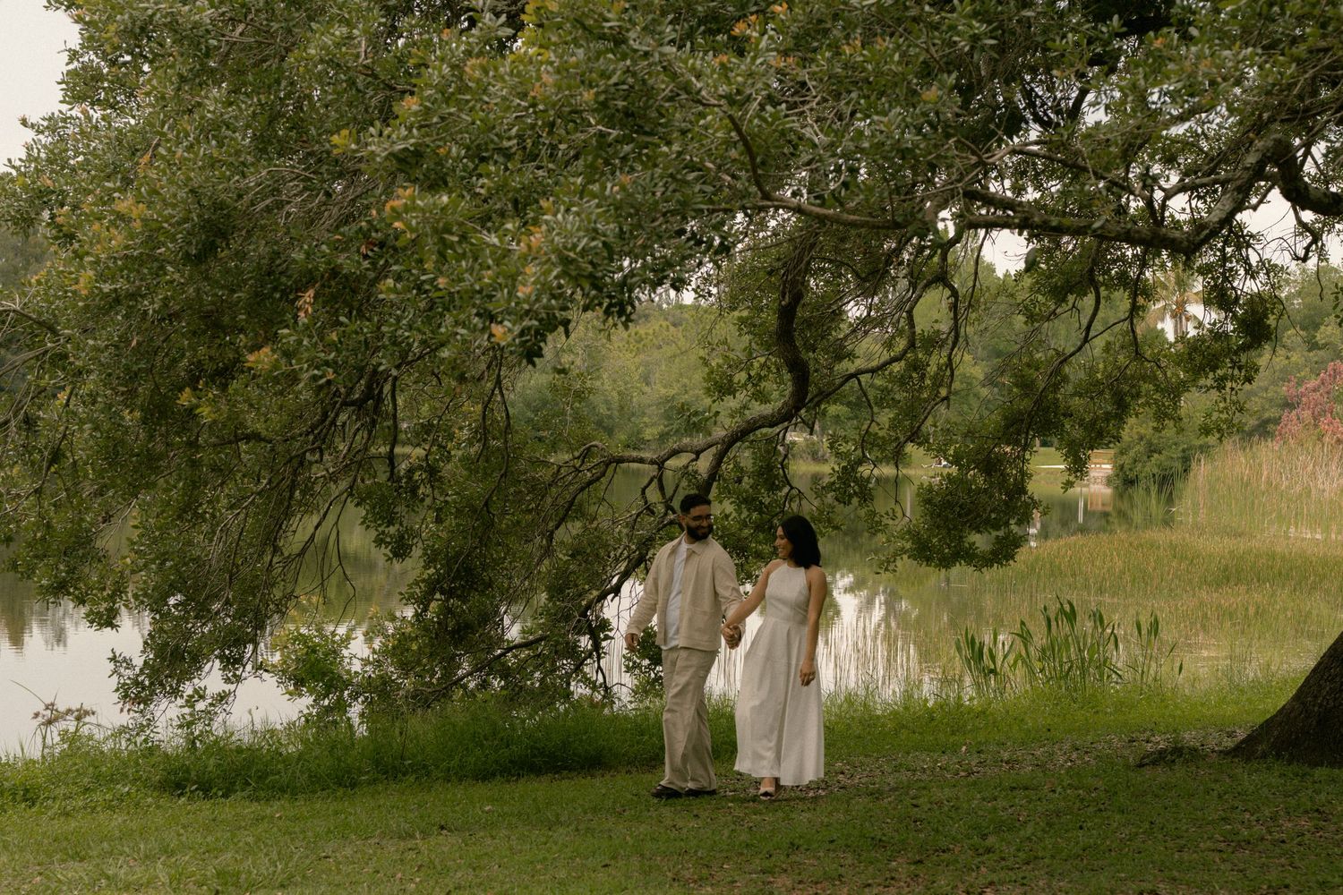 Couple in white attire holding hands near a serene lake under a large tree in a lush green landscape.