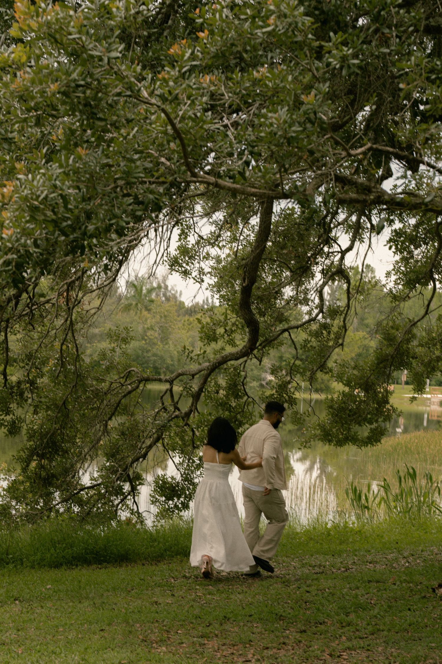 Couple walking together near a tranquil lake under sprawling tree branches in nature.
