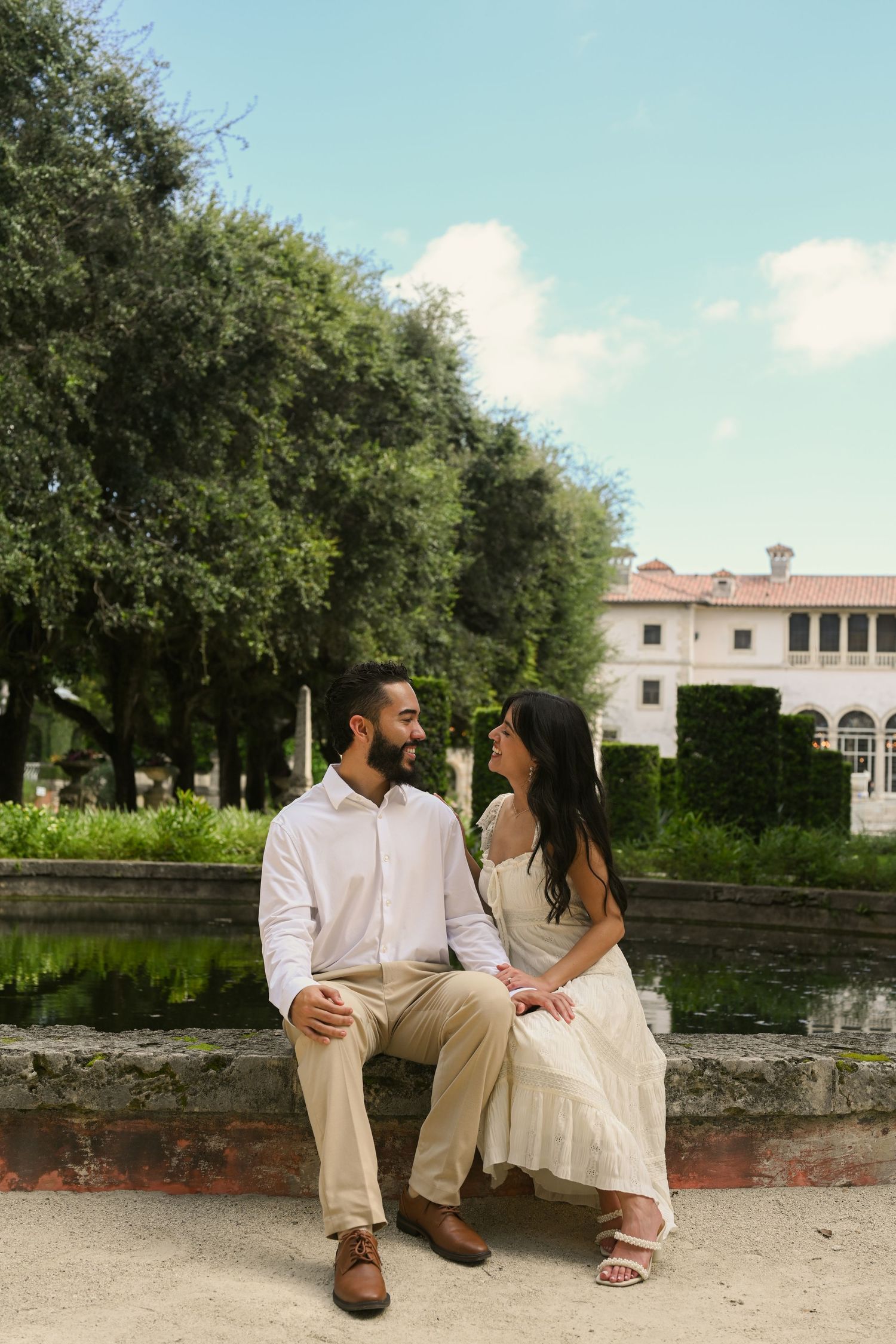 A couple in light summer wedding attire sits together on stone steps under shady trees.