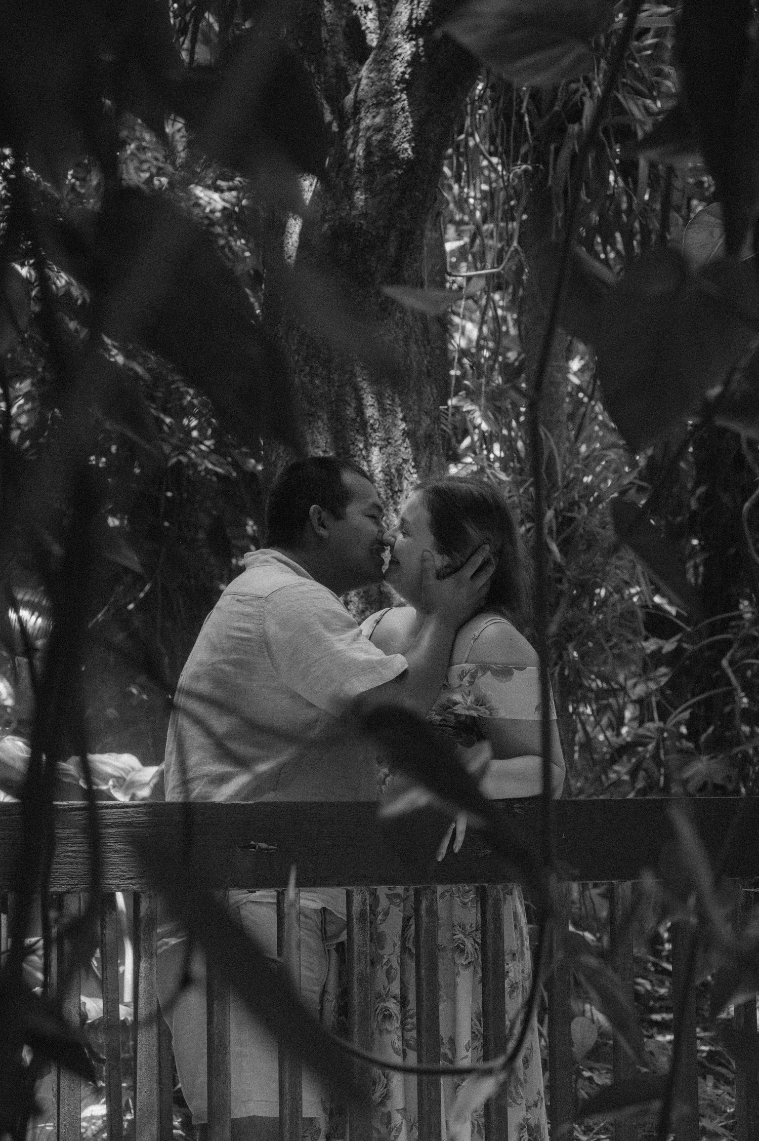 Black and white photo of a couple sharing a romantic kiss on a balcony surrounded by lush tropical trees.