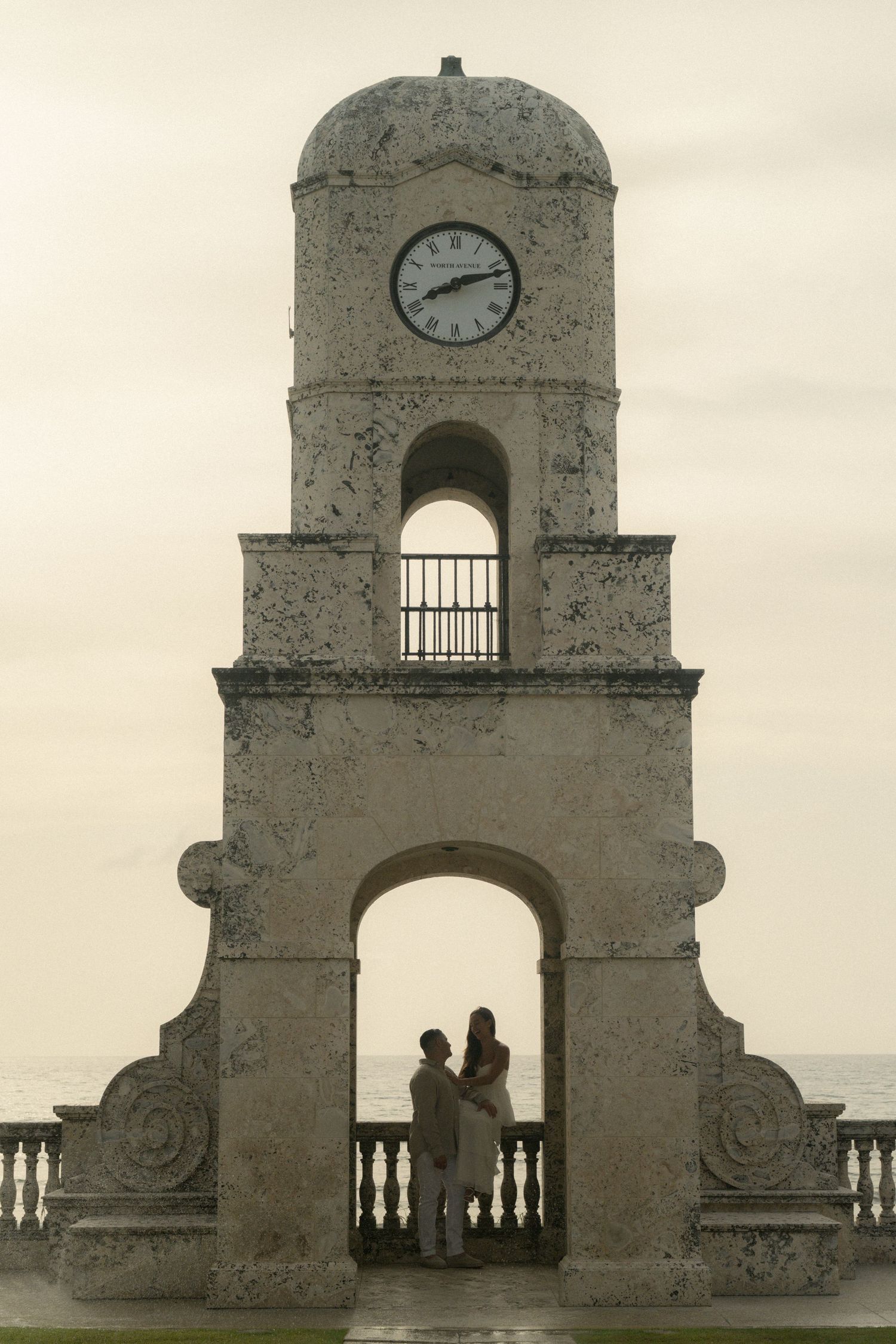 Couple embracing under historic clock tower arch with ocean backdrop at dusk.