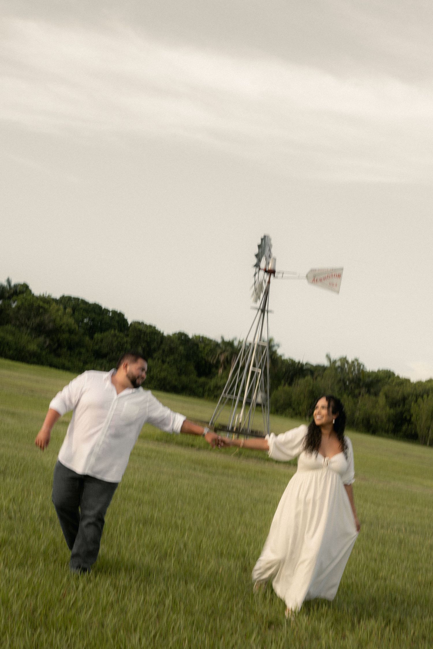 A couple holds hands and walks through a green field with a windmill in the background on a cloudy day.