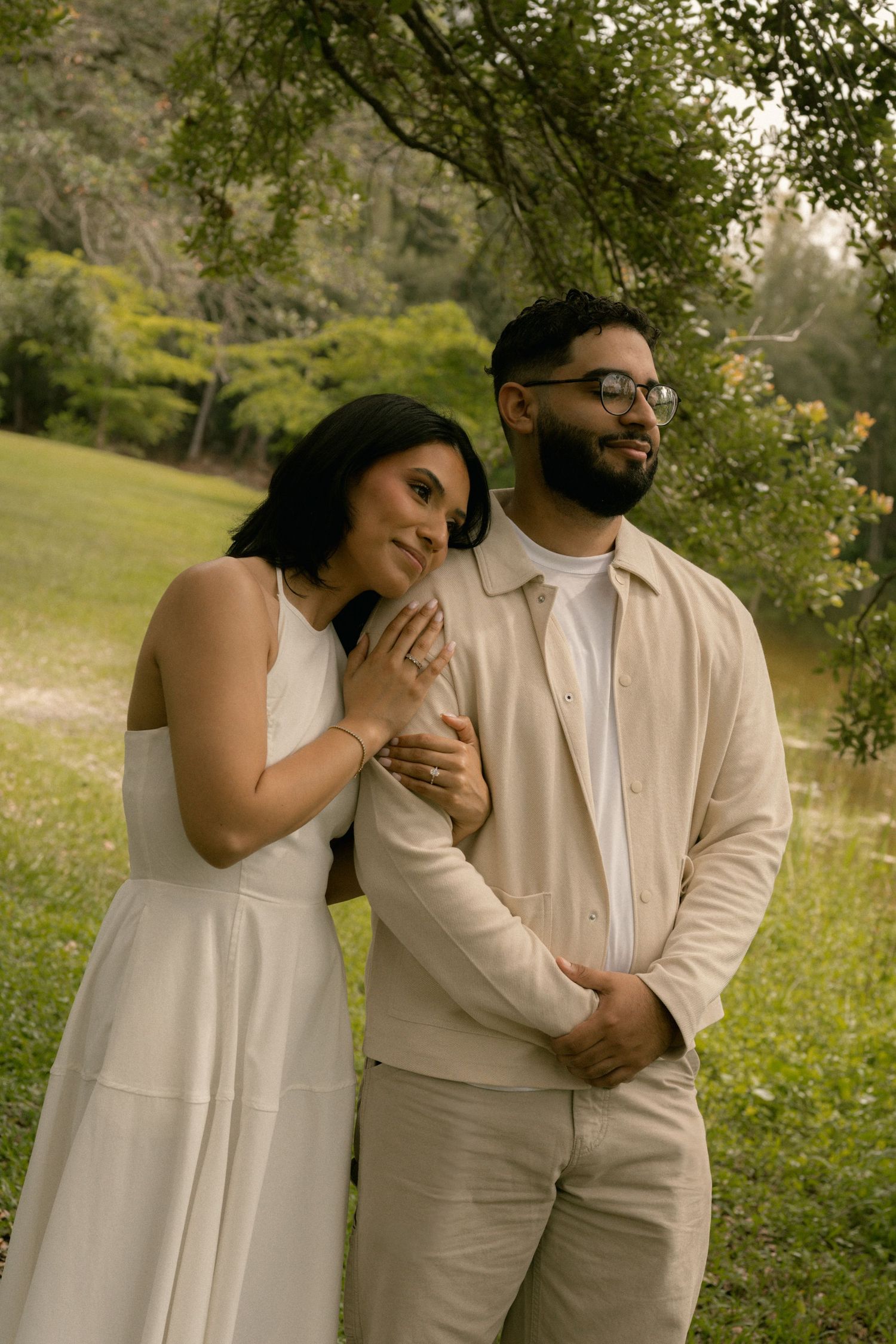 Elegant couple in white and beige outfits posing together in a lush green outdoor setting.