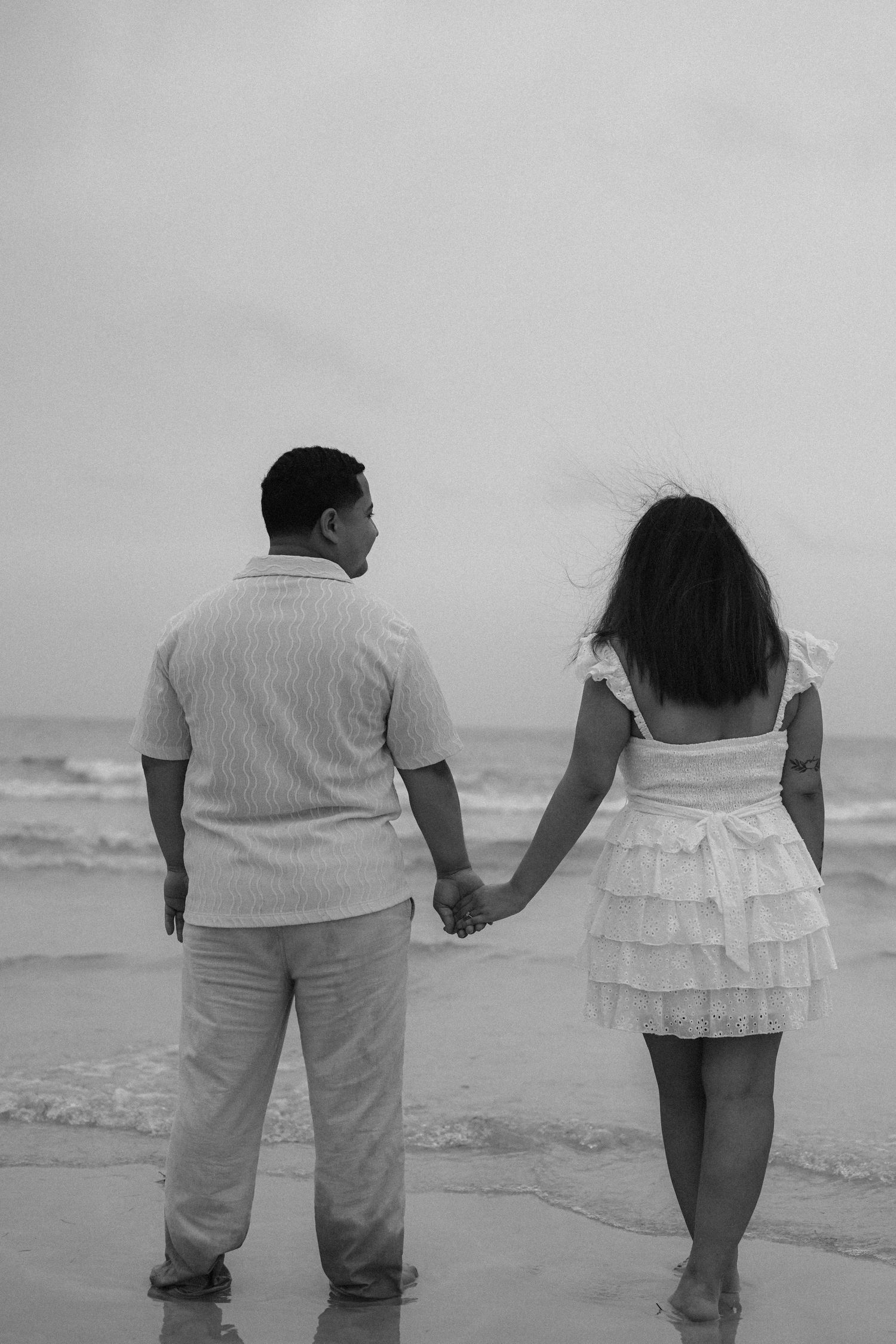 A couple in white attire walking hand in hand along the ocean's edge.