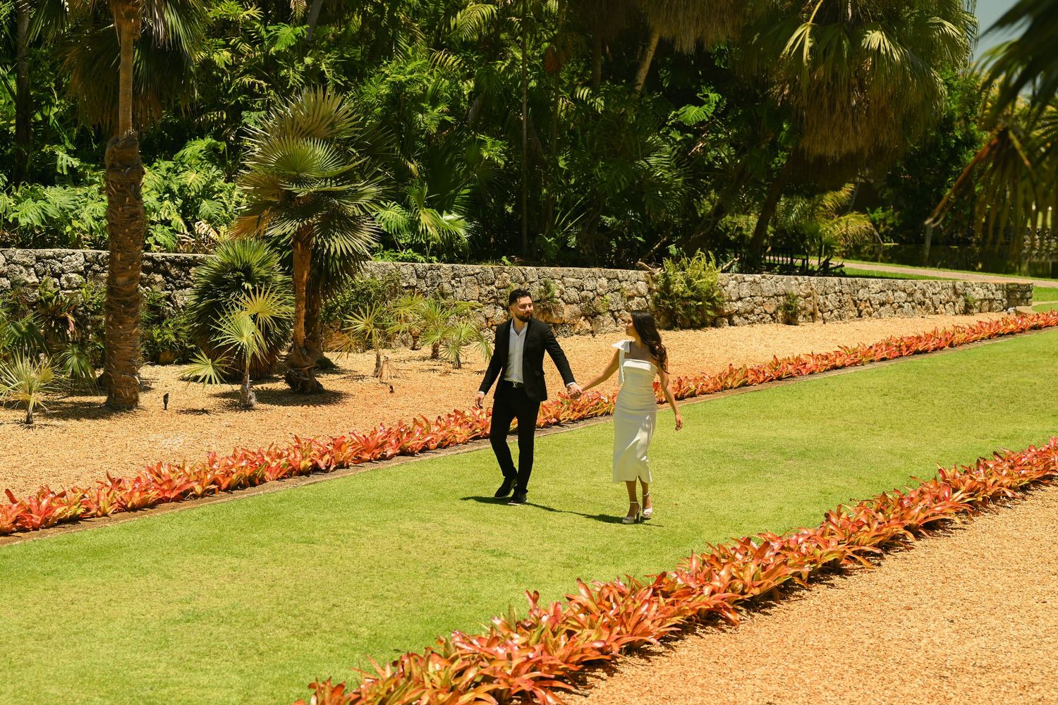 A couple walks along a winding garden path bordered by bright coral-colored flowers.