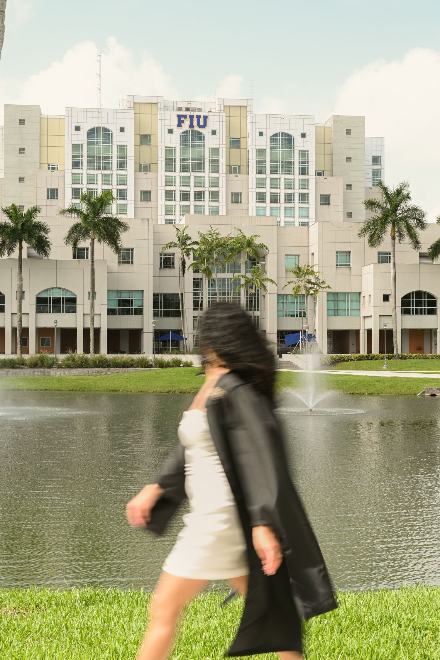 Campus building reflected in lake with palm trees and graduate in foreground.