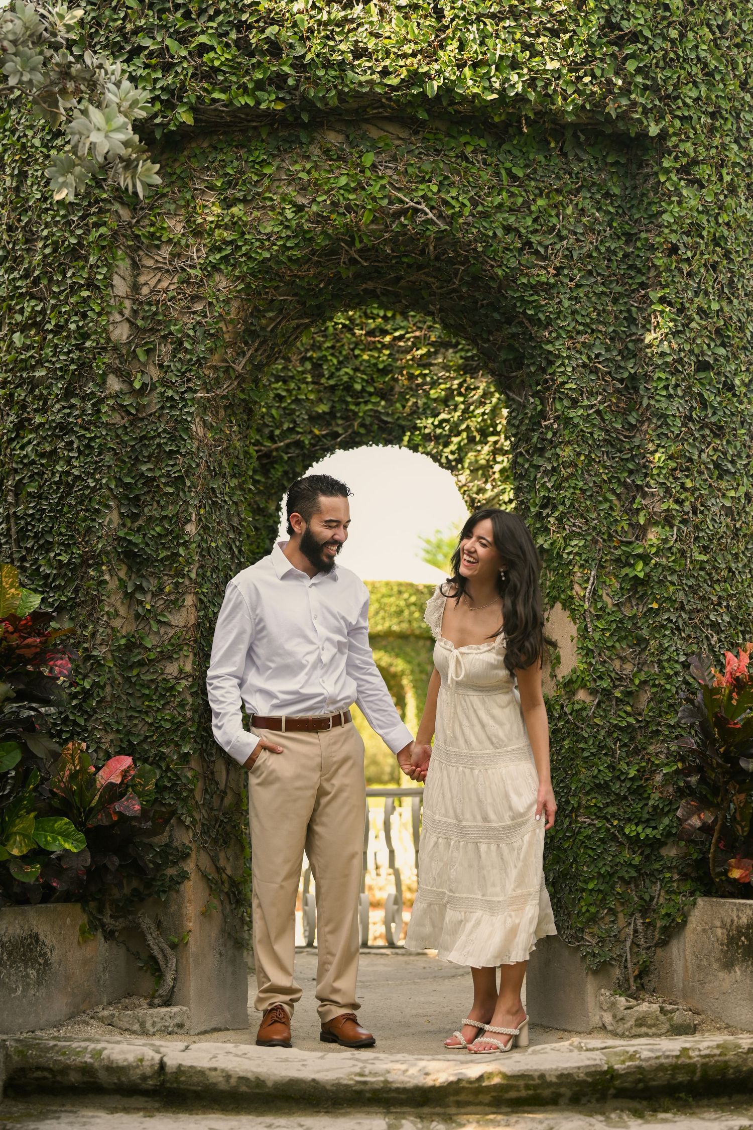 A couple shares a romantic moment in a lush garden setting with stone archways and greenery.
