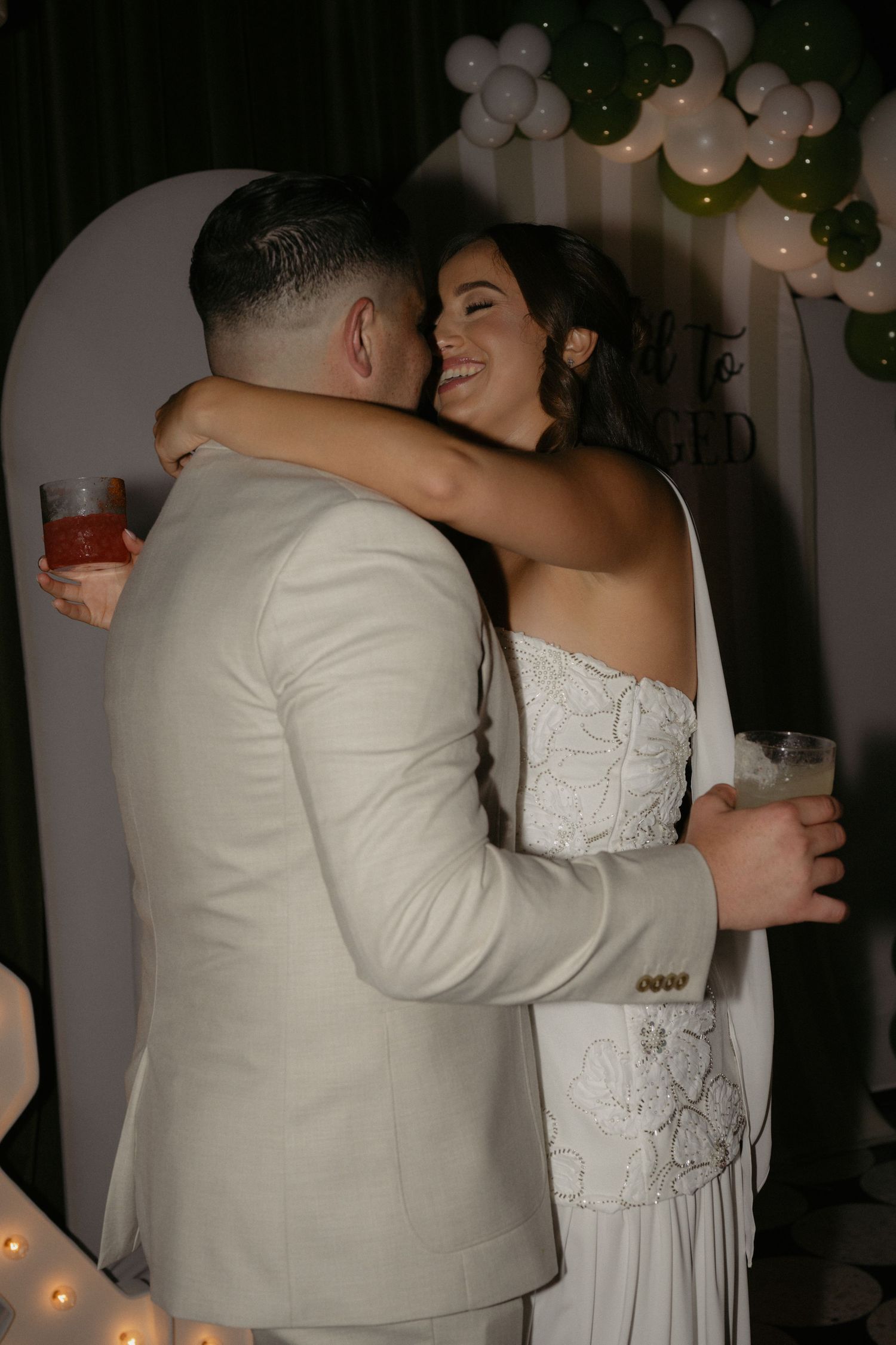 Bride and groom embrace and smile holding drinks at wedding reception with green and white balloon arch.