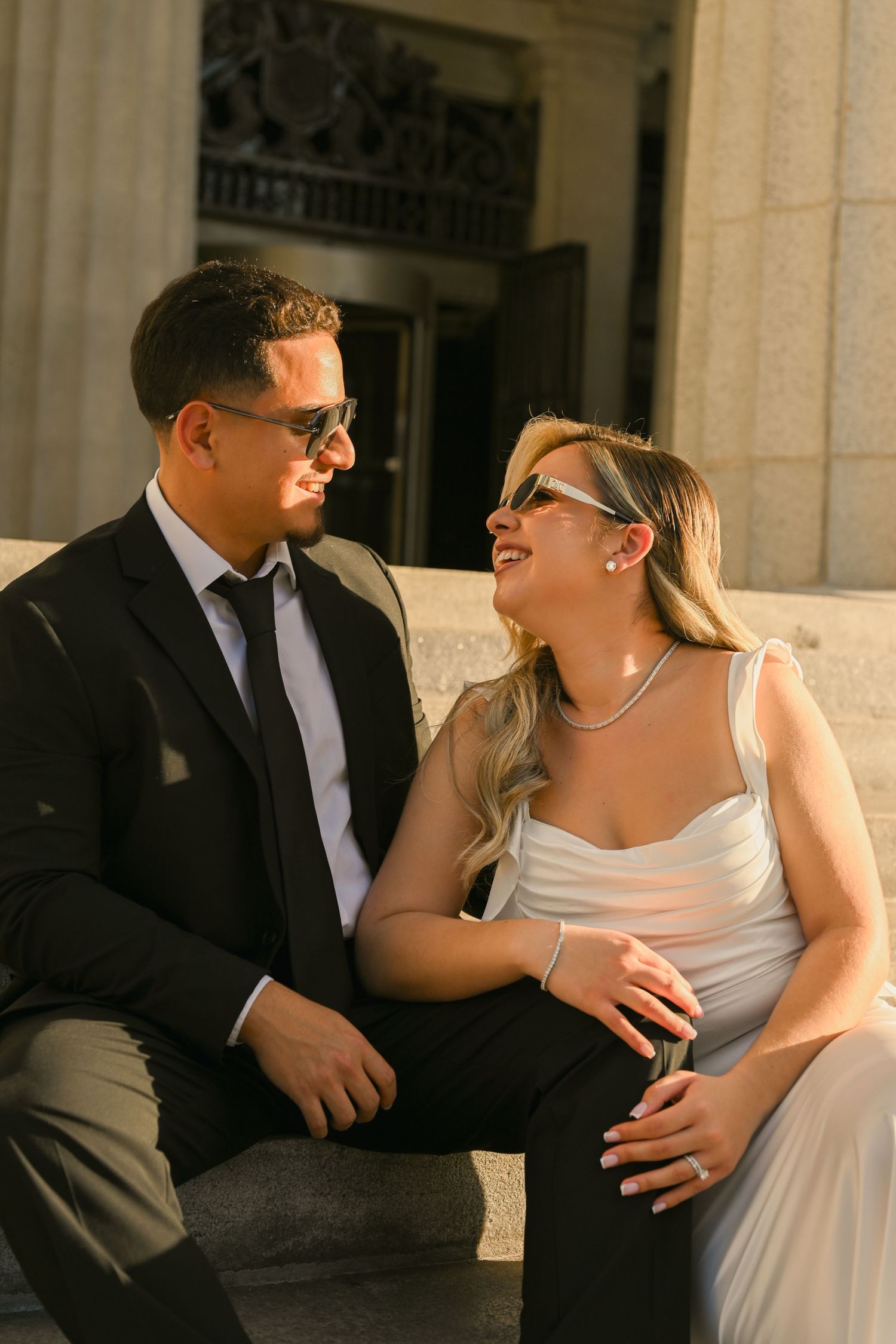Couple in formal attire sitting on stone steps, smiling at each other in golden sunlight.
